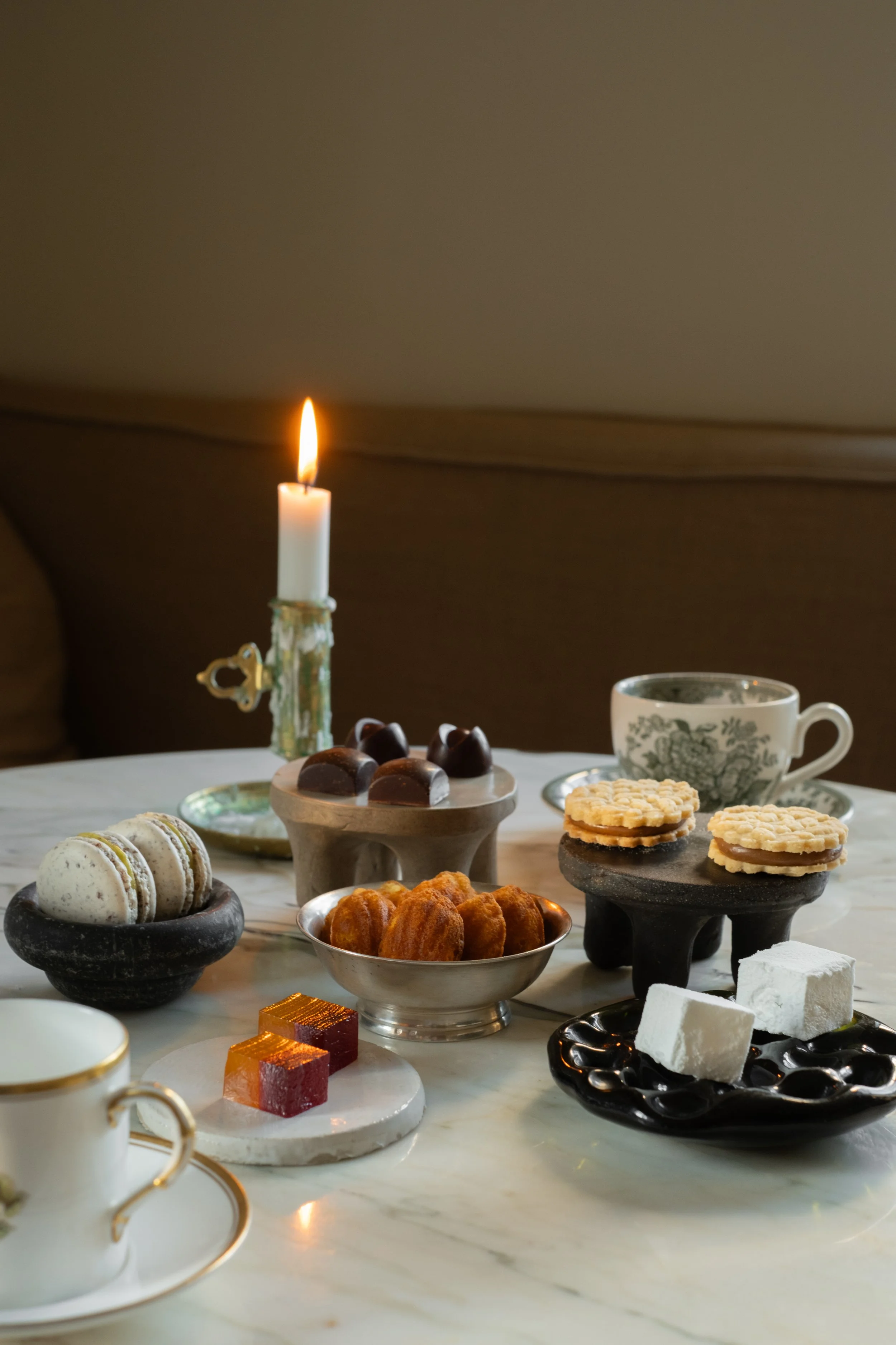 Tekopp med te, kakefat med sjokolade, makroner, småkaker, marshmallows og gelégodteri på et bord med lysende stearinlys.