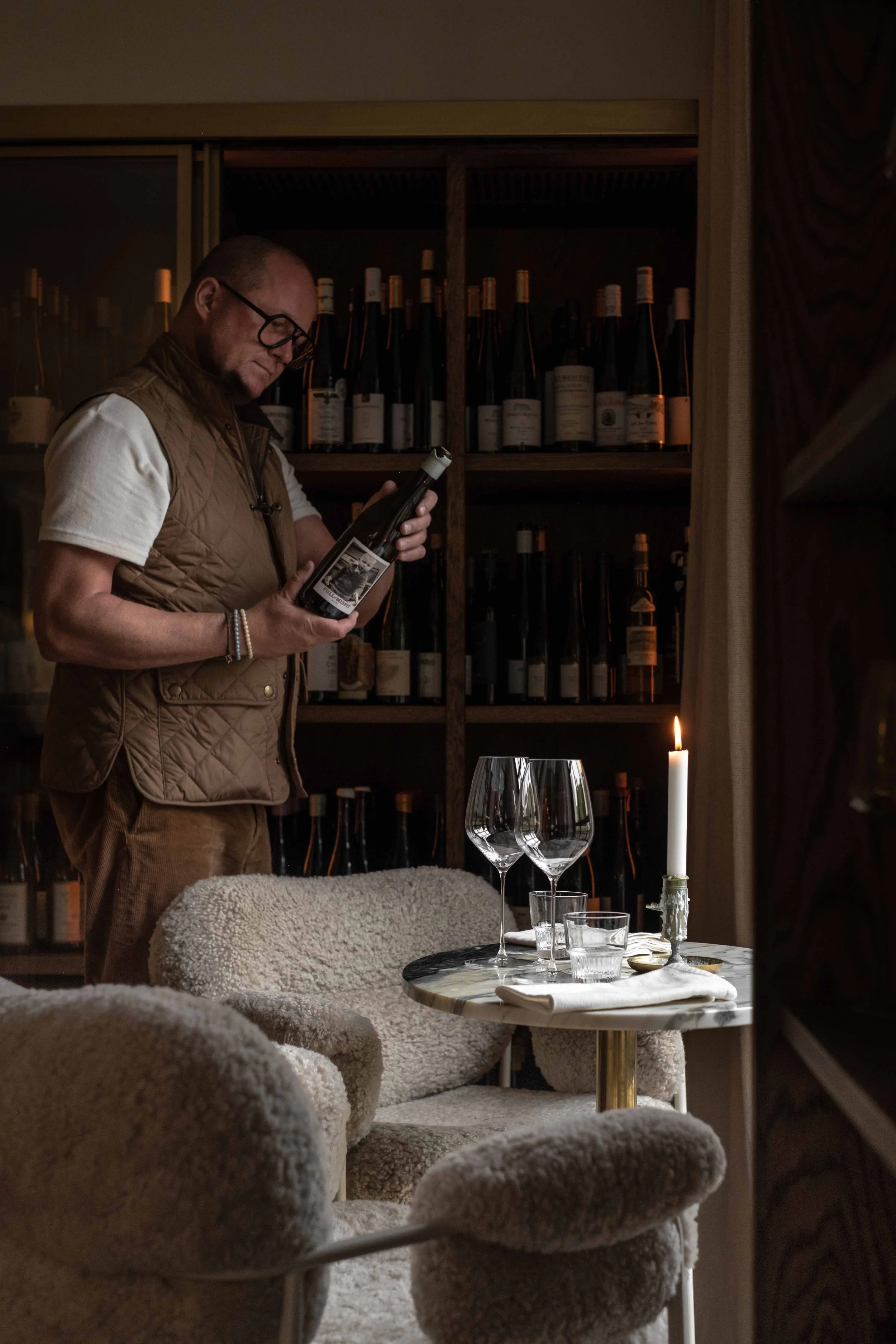 En mann som står ved et bord med to store vinglass og en lysende candle. Han ser på en vinflaske, mens det er et rekke av vinflasker på hyllen bak ham.