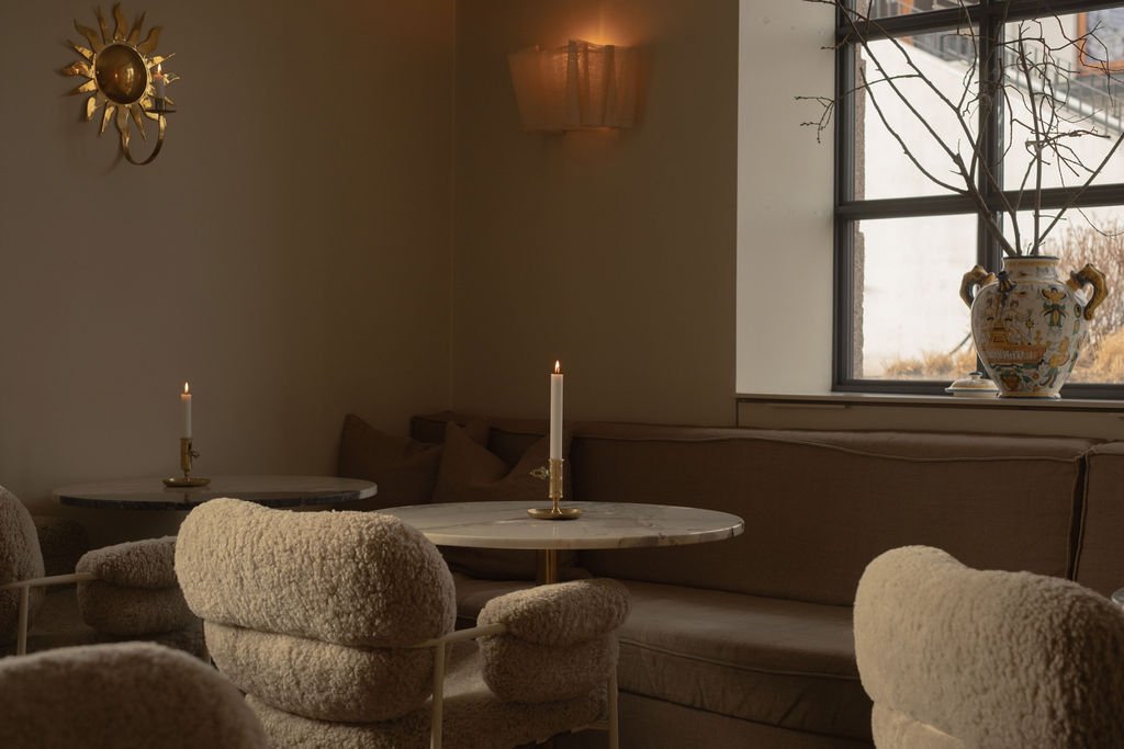 Interiør av en koselig stue med beige møbler, to bord med lys og en stor vase med grener på vinduskarmen.