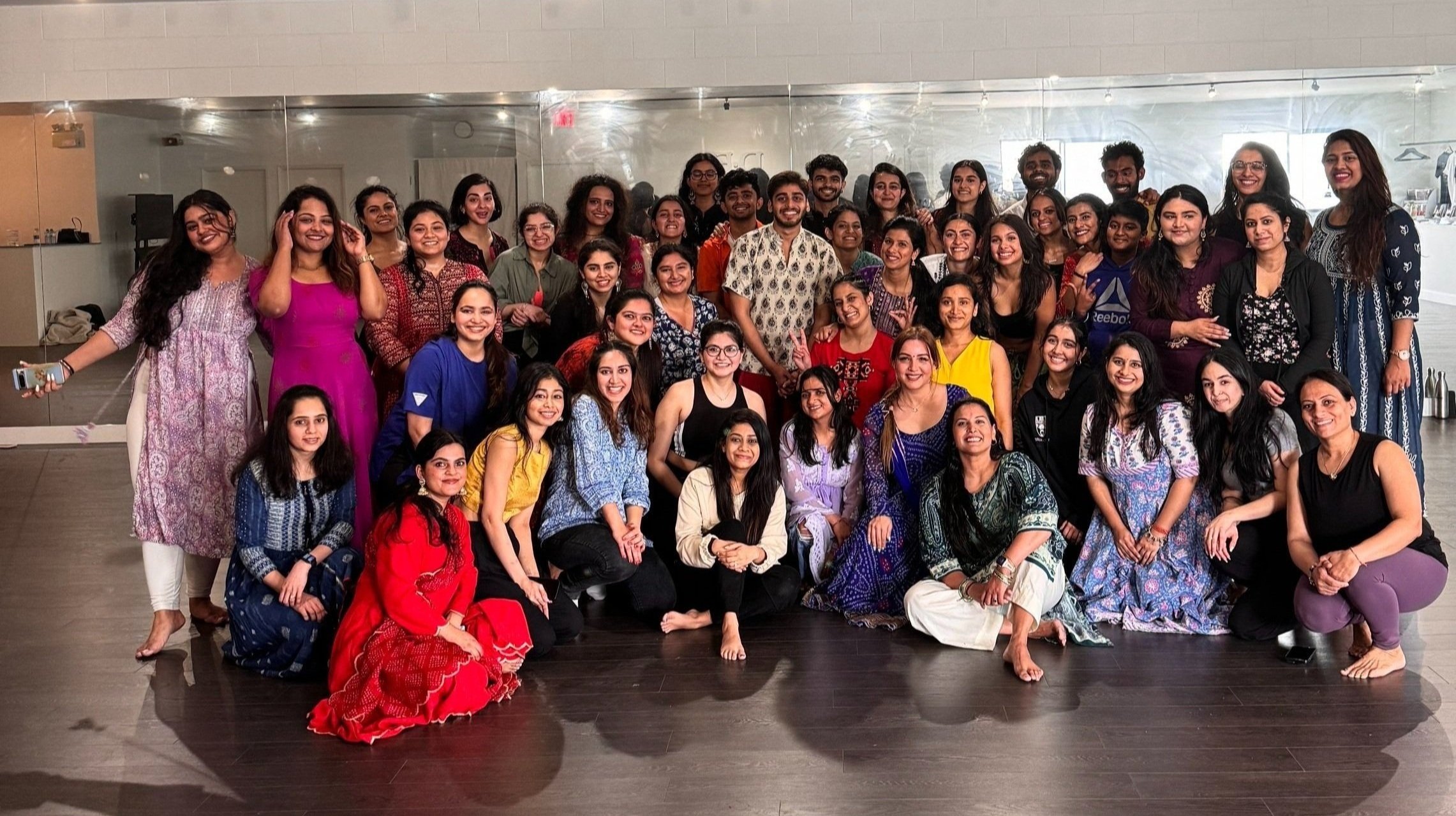 Group of smiling women and men in colorful traditional and casual clothing posing indoors for a group photo.