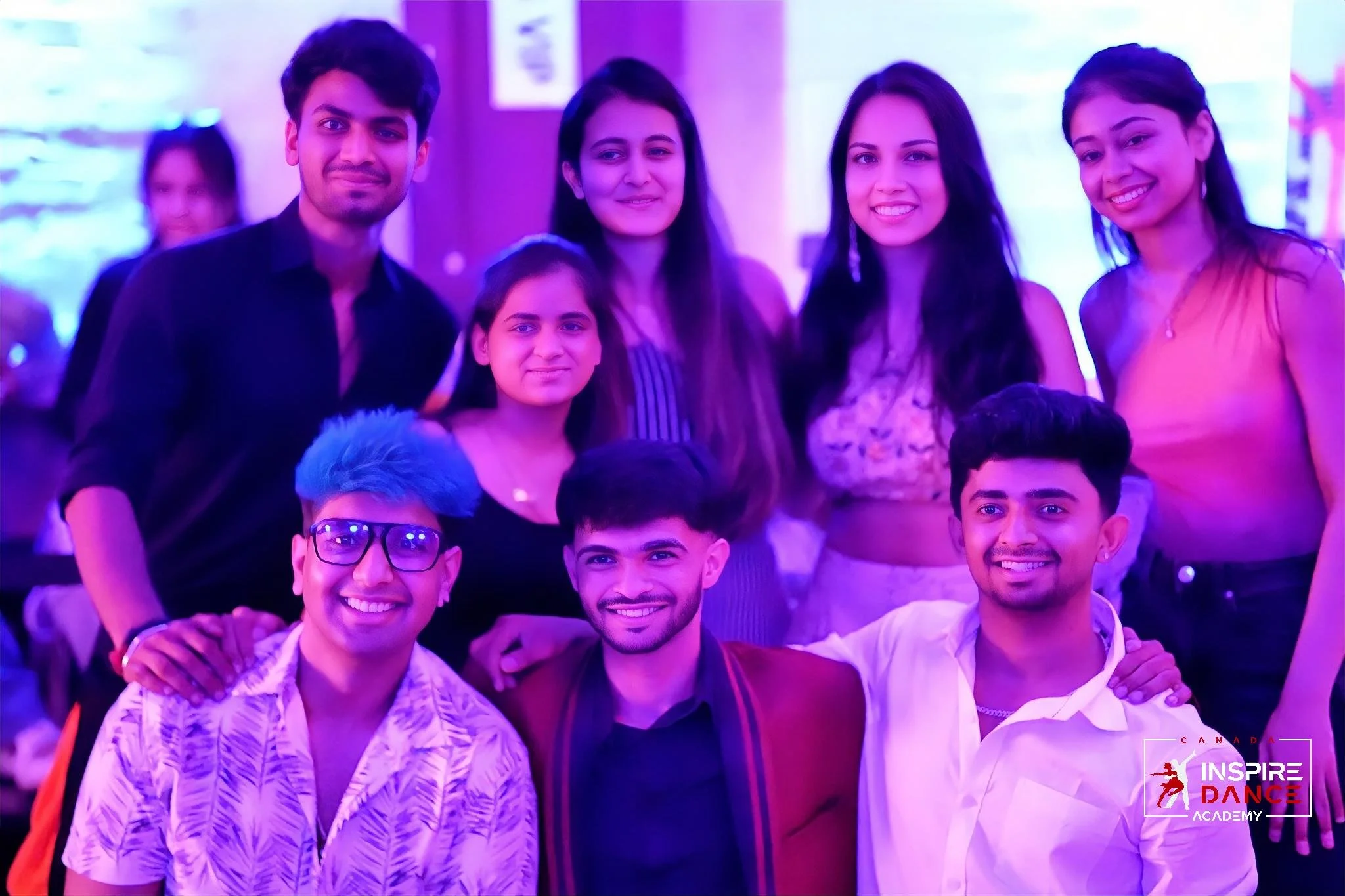 Group of nine people posing together at a dance event with blue and purple lighting, some smiling, some with serious expressions, in a social setting.