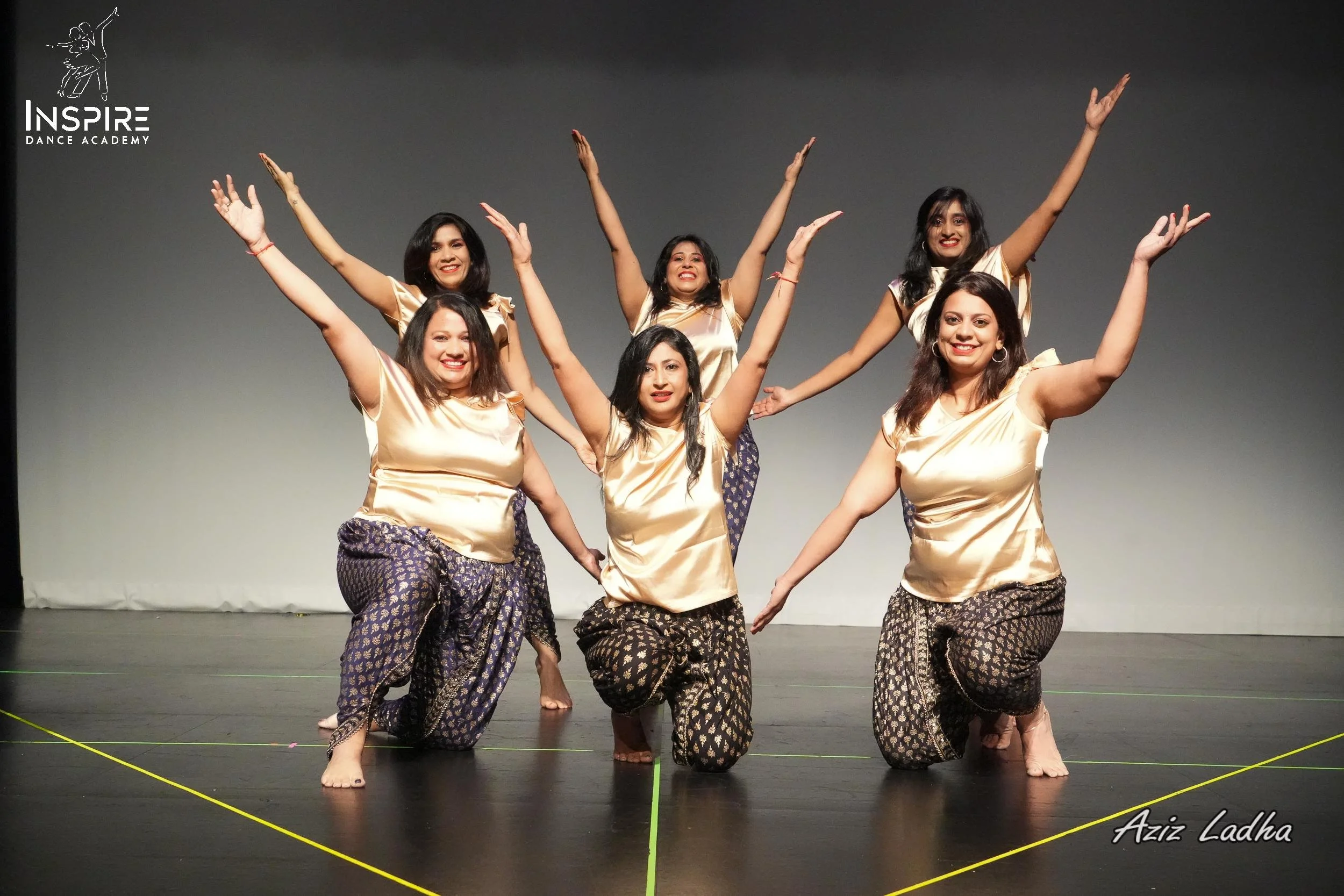 Six women in traditional clothing performing a dance on stage, with arms raised and smiling, in front of a plain background at Inspire Dance Academy.