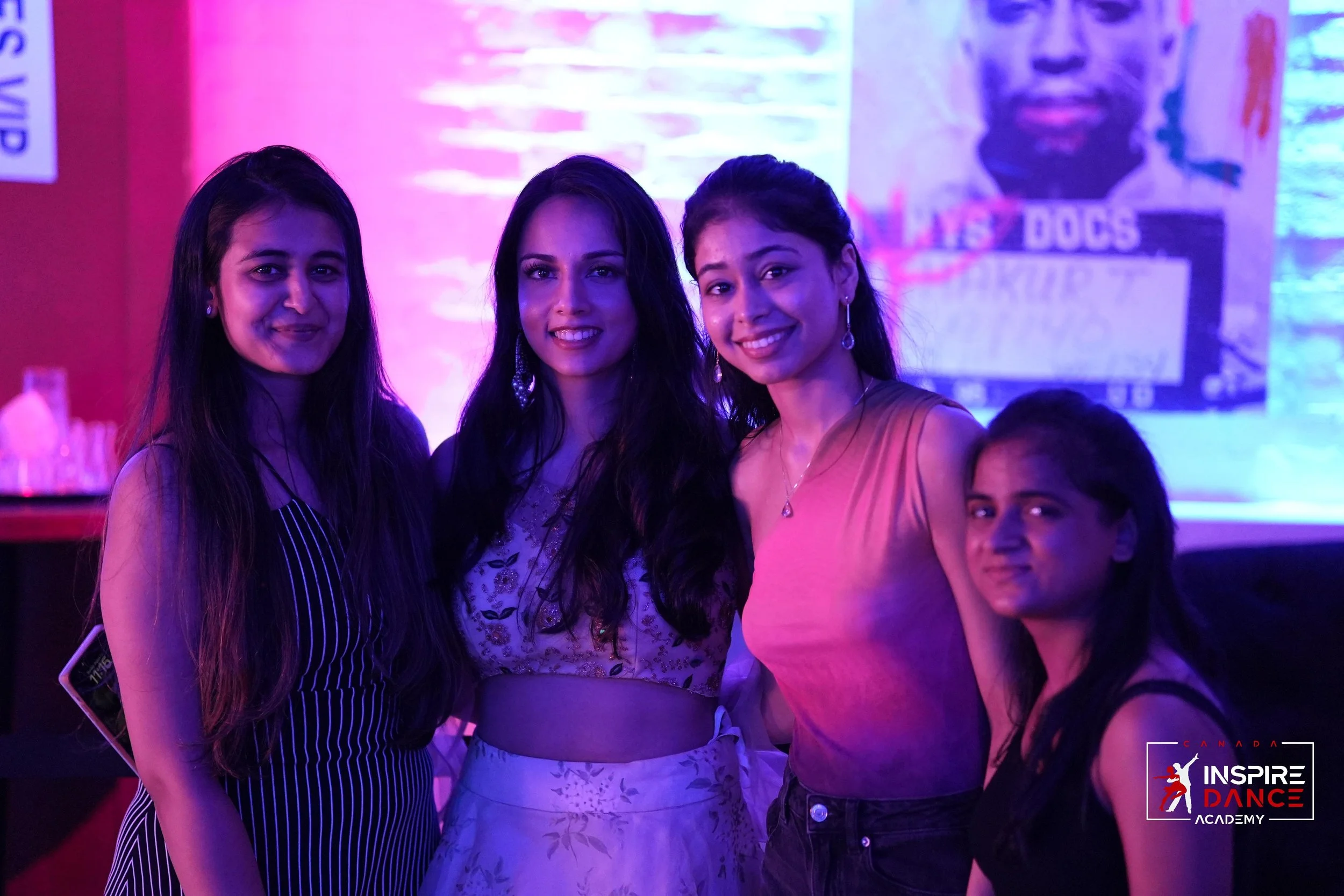 Four women posing together indoors, with colorful lighting and posters in the background, at Inspire Dance Academy event.