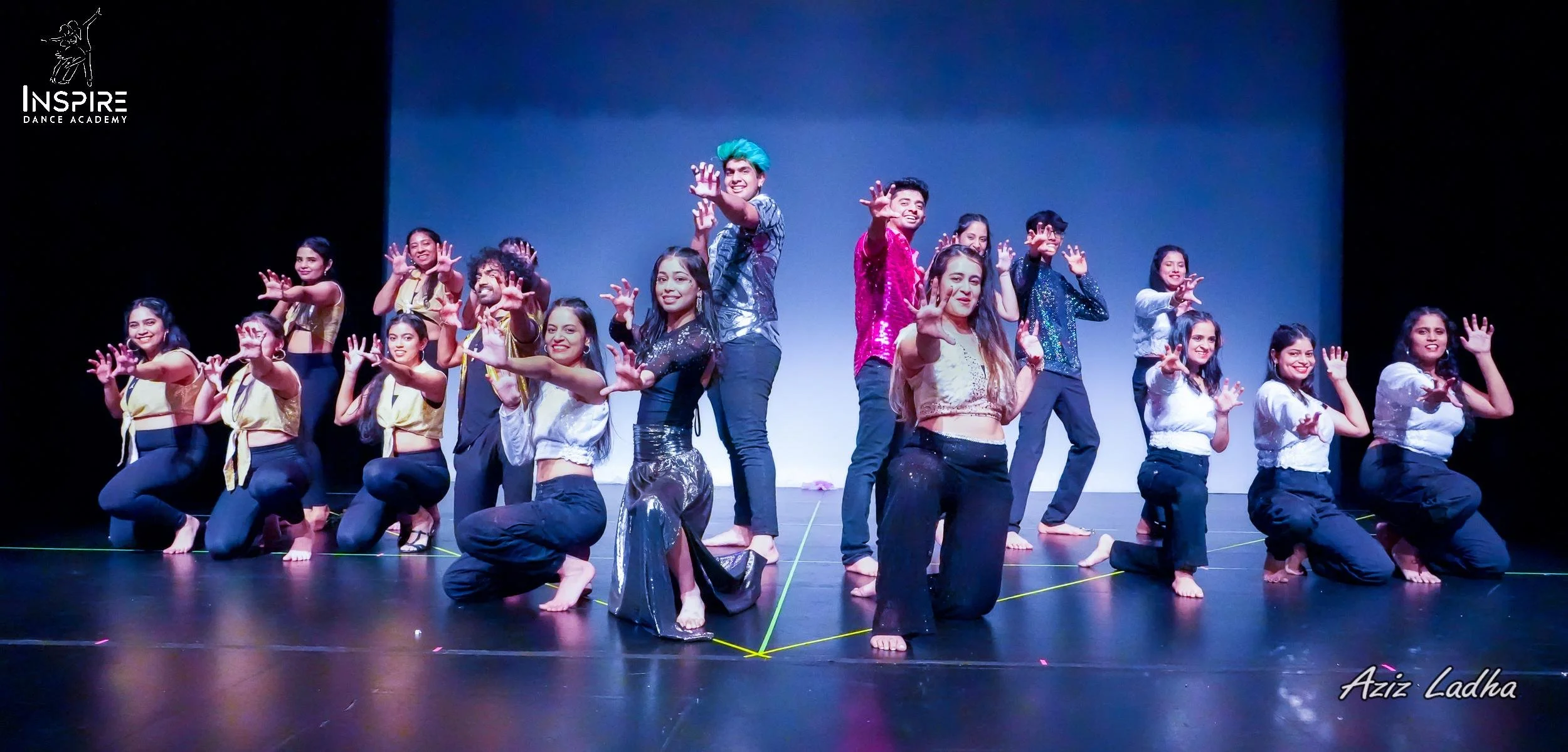 Group of young dancers on stage, posing with hands reaching forward, smiling, wearing colorful outfits, performing at Inspire Dance Academy.