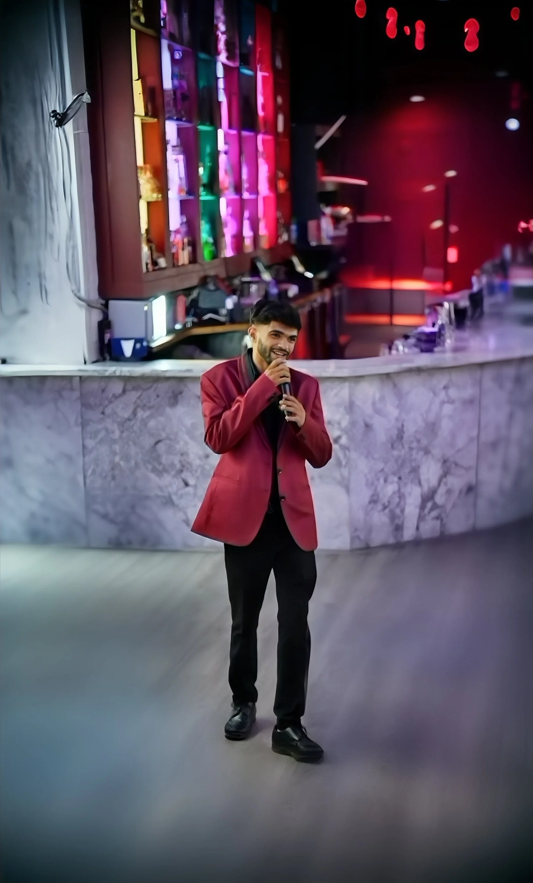 A man in a red blazer and black pants standing at a bar counter, holding a microphone and smiling, with colorful illuminated shelves behind him in a dimly lit venue.