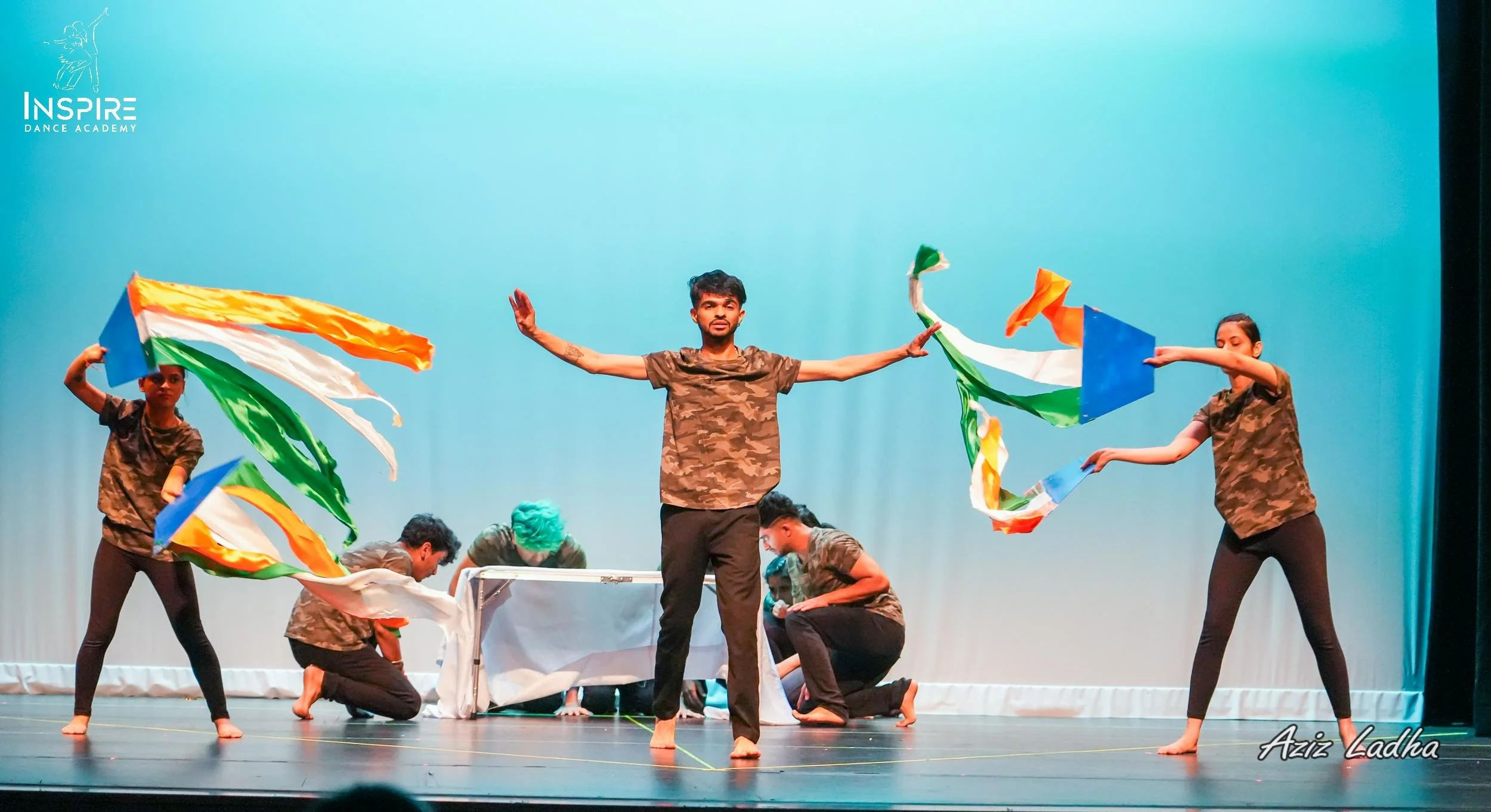 Performers dressed in camouflage shirts and black pants on stage, with some holding colorful fabric flags resembling the Indian flag, during a dance or theatrical performance at Inspire Dance Academy.