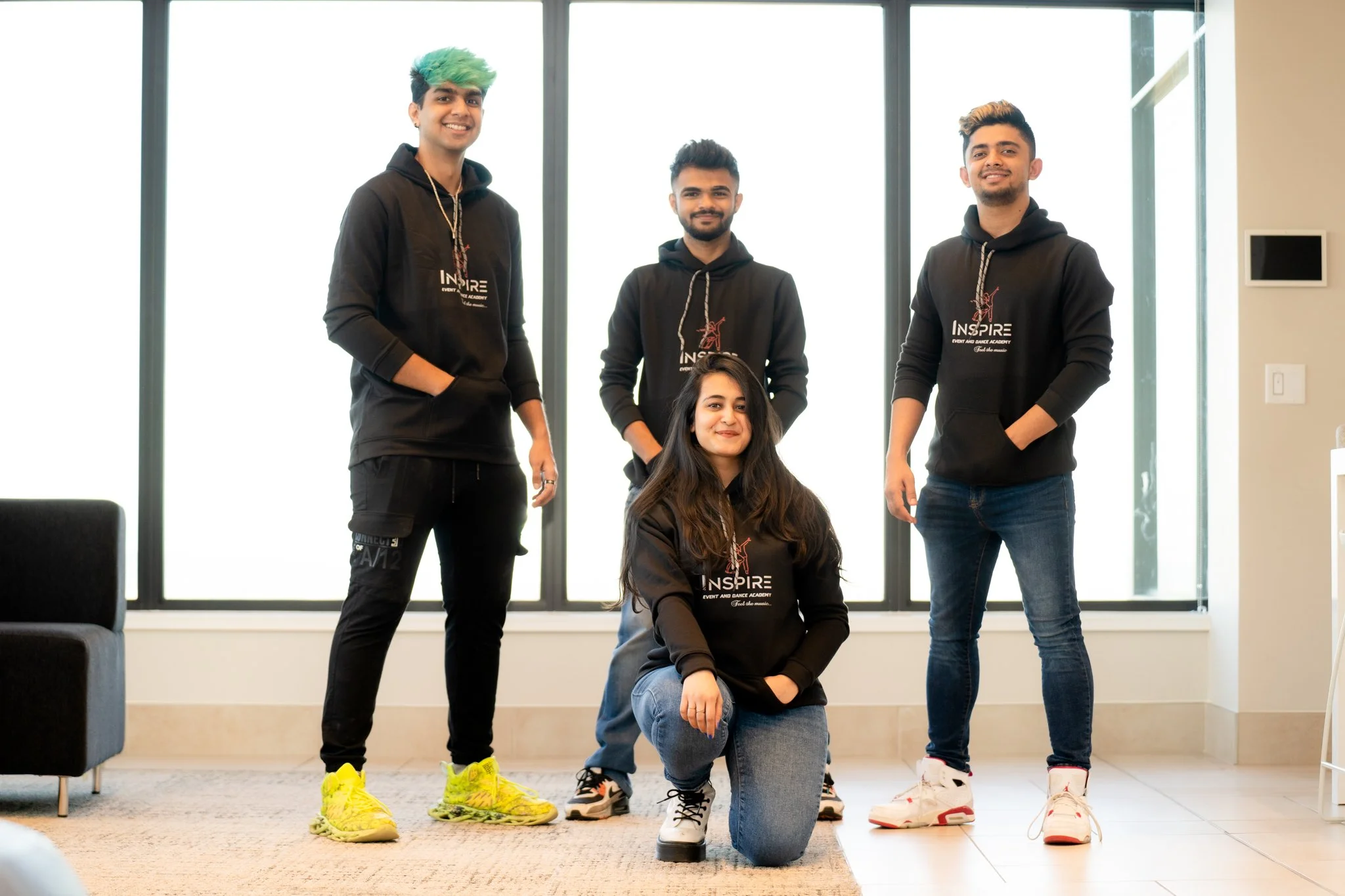 Four young adults, one woman kneeling in front and three men standing behind her, all wearing black hoodies with a dance logo, inside a room with large windows.
