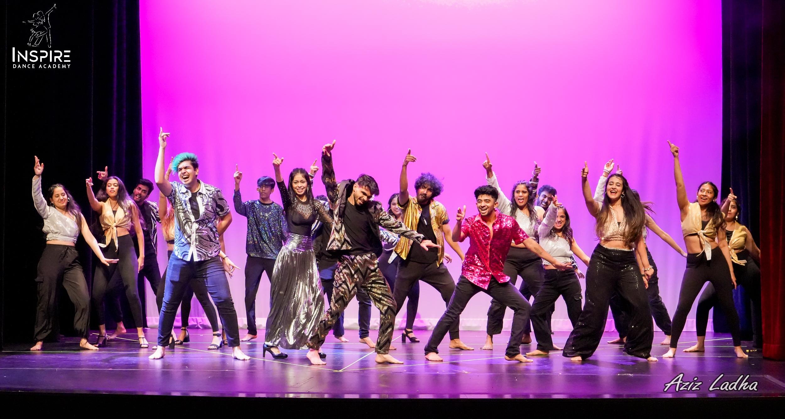 Group of diverse young dancers performing on stage with a pink backdrop, smiling and dancing energetically.