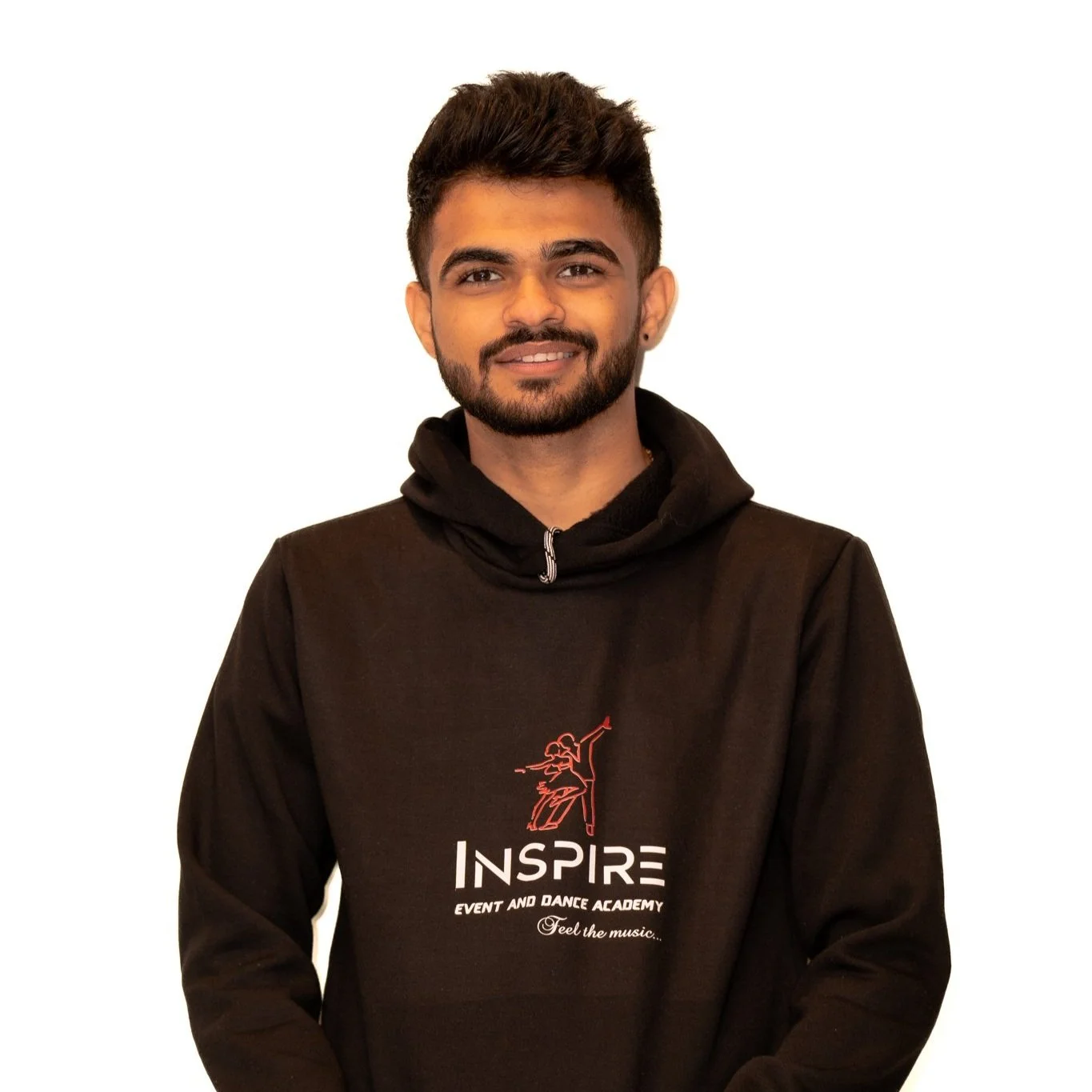 A young man with dark hair and a beard smiling, wearing a black hoodie with an 'Inspire' logo and text that reads 'Event and Dance Academy, Feel the music...' against a plain white background.