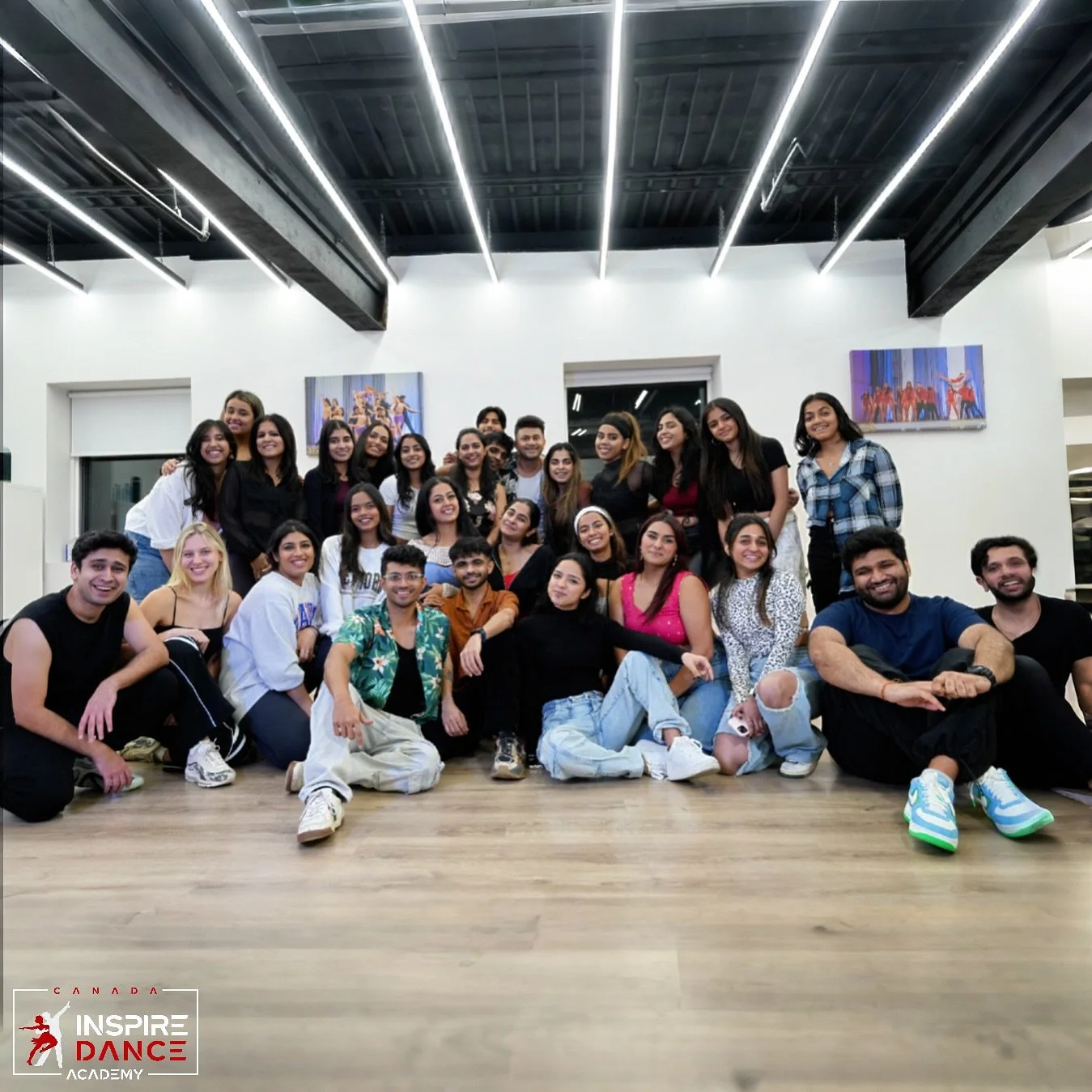 Group of young adults posing together indoors, some sitting, some standing, smiling at the camera, with a modern dance studio background including artwork on the walls.