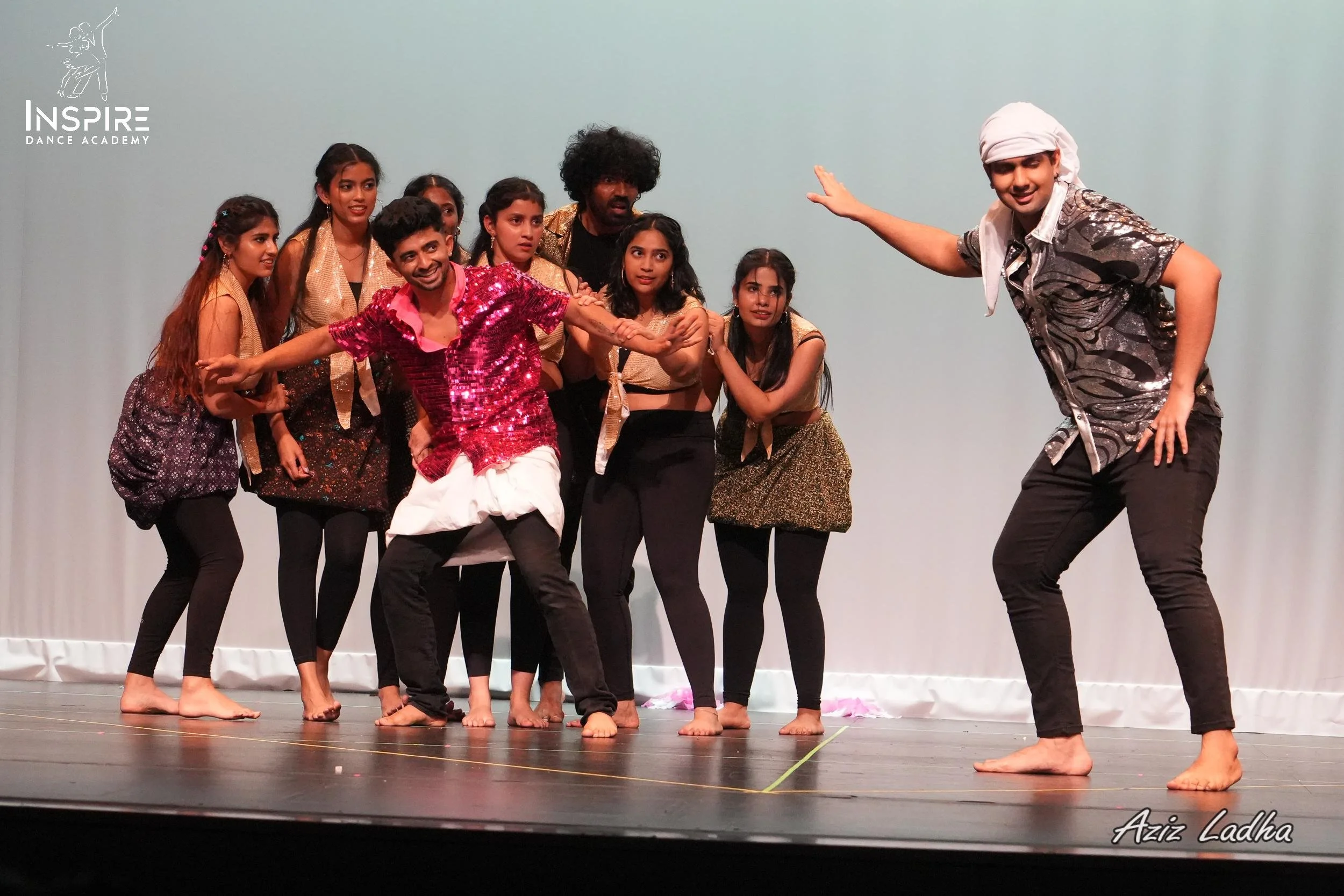Group of young performers on stage, some with expressive poses, participating in a theatrical or dance performance at Inspire Dance Academy. The stage is plain white backdrop, and two male performers are in front, one in a shiny shirt and the other in a pink sequined shirt, with several female performers in the background.