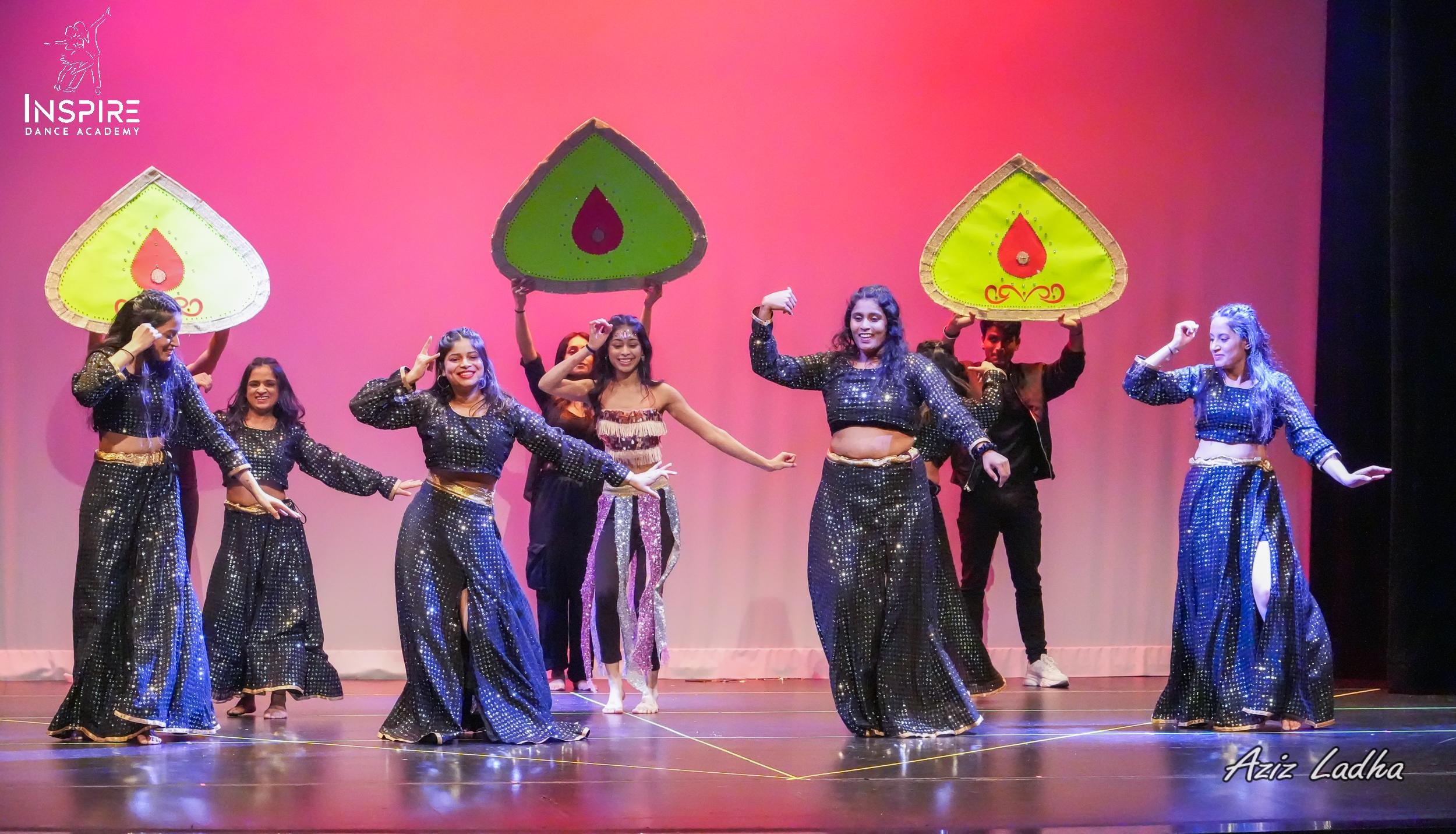 Group of young women performing a dance on stage, some holding large colorful umbrellas and dressed in black and sparkly outfits, with a pink and purple background.