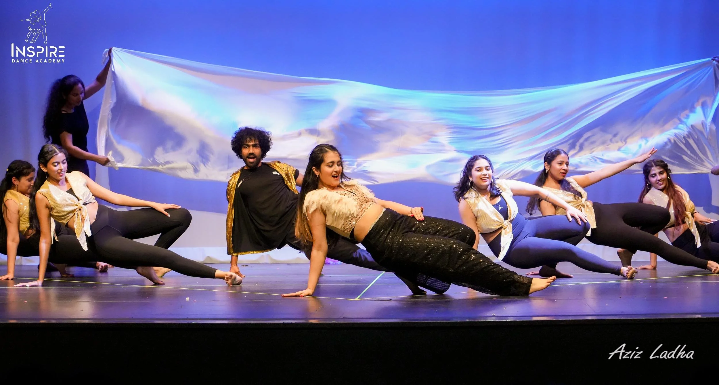 Dance performers on stage, holding a large fabric, during a dance performance at Inspire Dance Academy, with a blue background and stage lighting.