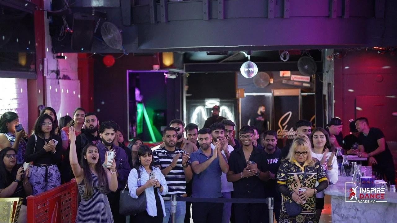 A group of people standing inside a dance studio or club, some holding phones, with colorful lighting and a bar area in the background.