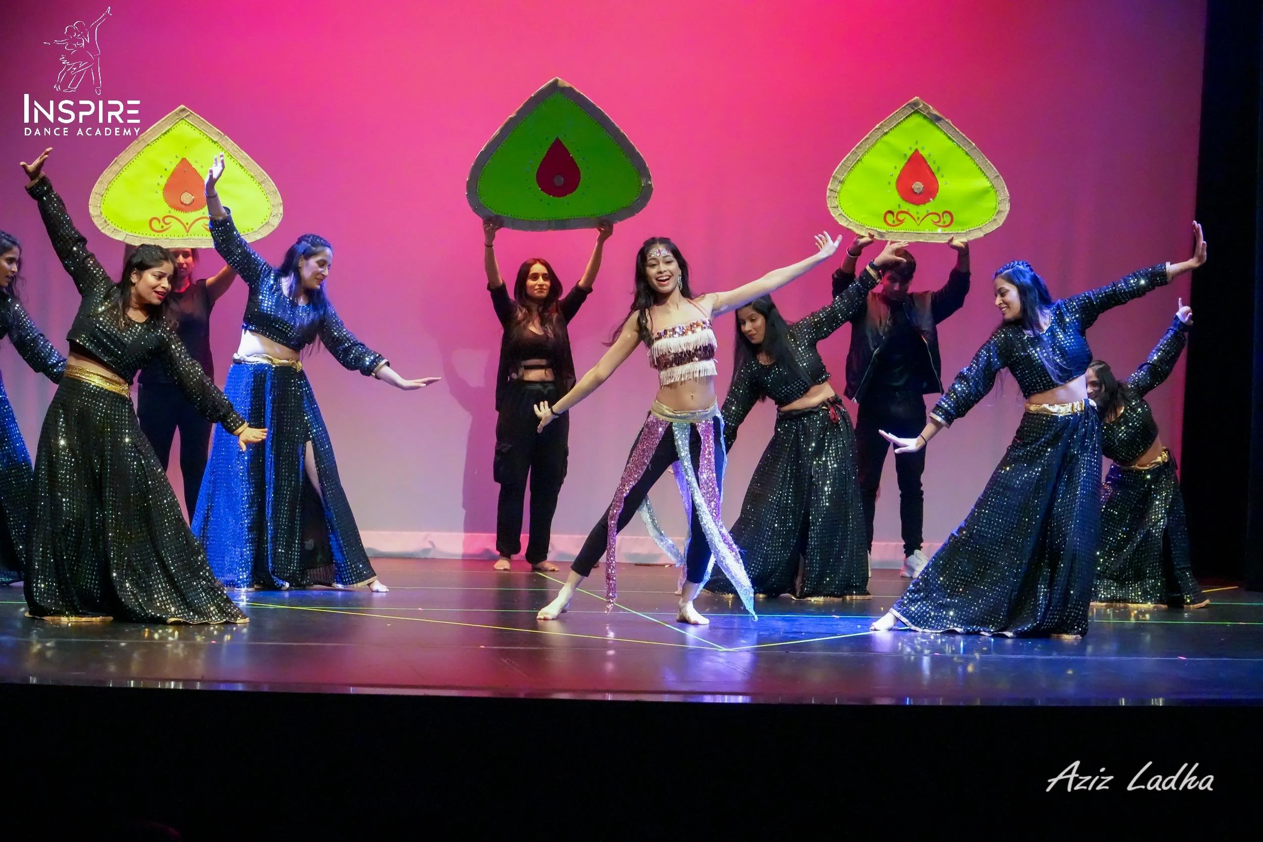 Group of dancers performing on stage with colorful backdrops, some holding decorative umbrellas, during a dance recital at Inspire Dance Academy.