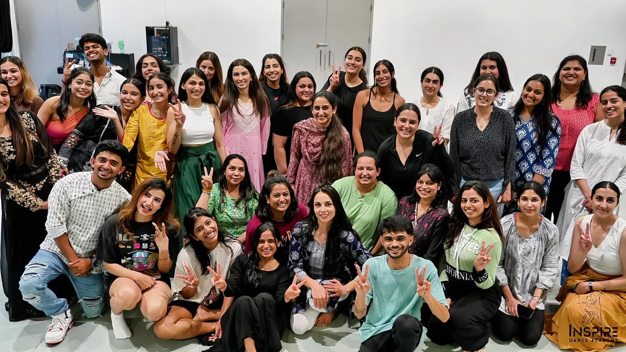 Group of young adults, including men and women of diverse backgrounds, smiling and posing together in a room. Some are standing, some squatting or kneeling in front, making peace signs, standing against a plain wall with doors and equipment in the background. The bottom right corner includes the logo for Inspire Dance Academy.