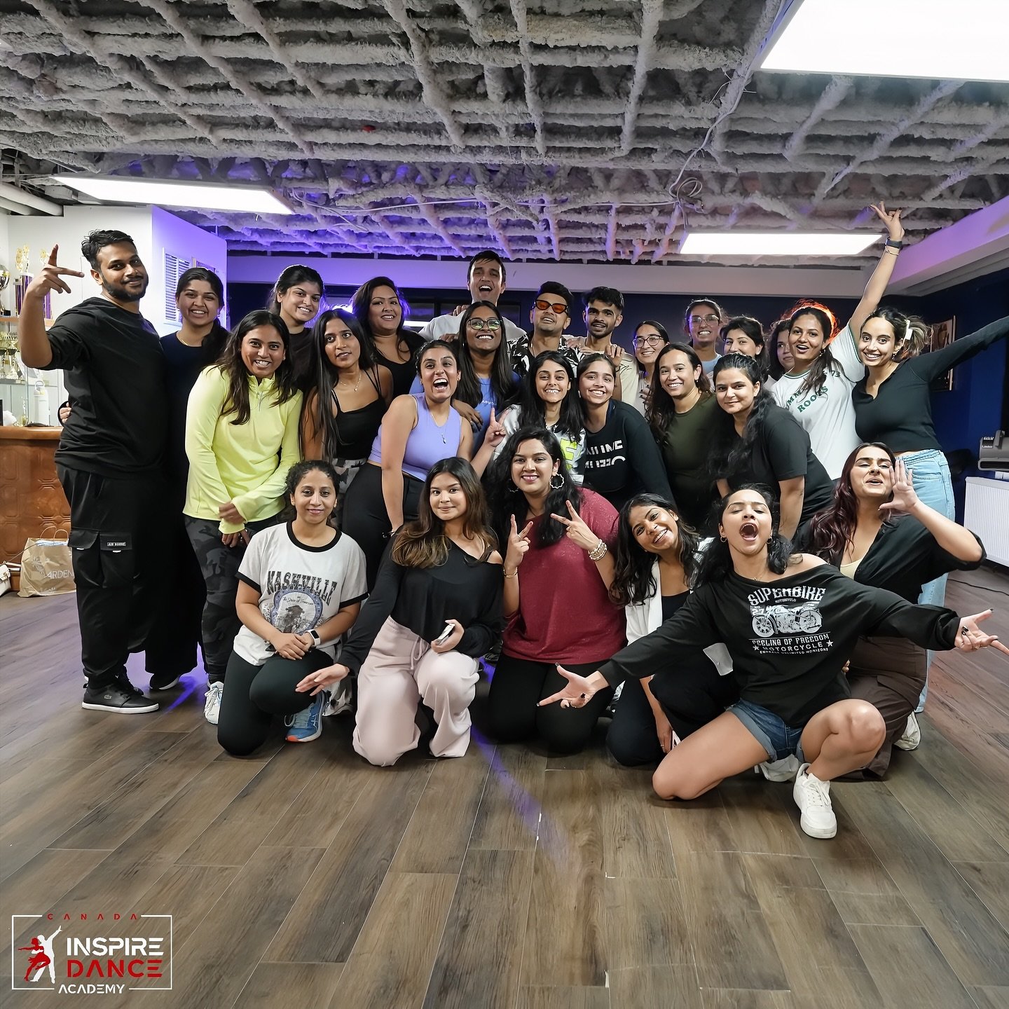 Group of young adults smiling, posing, and celebrating in a dance studio with purple lighting, wooden floor, and a ceiling under construction. Inspire Dance Academy logo is visible in the bottom left corner.