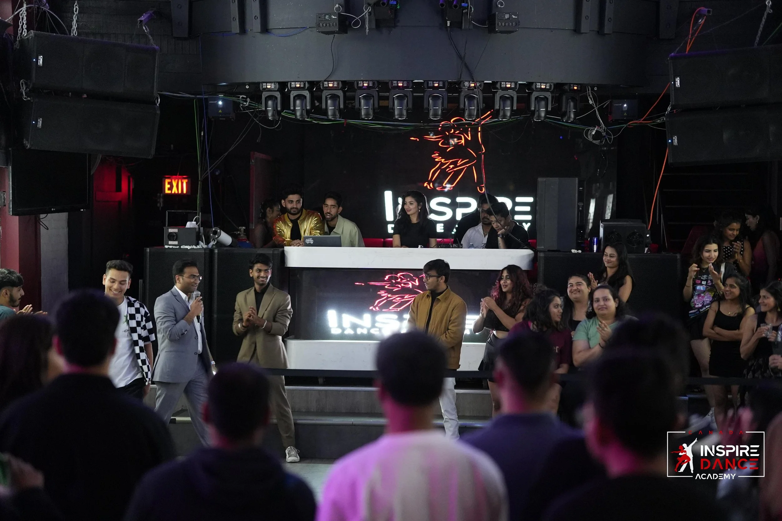 Dance event at Inspire Dance Academy with people gathered in front of stage, a DJ booth in the background, and neon signs on the wall.