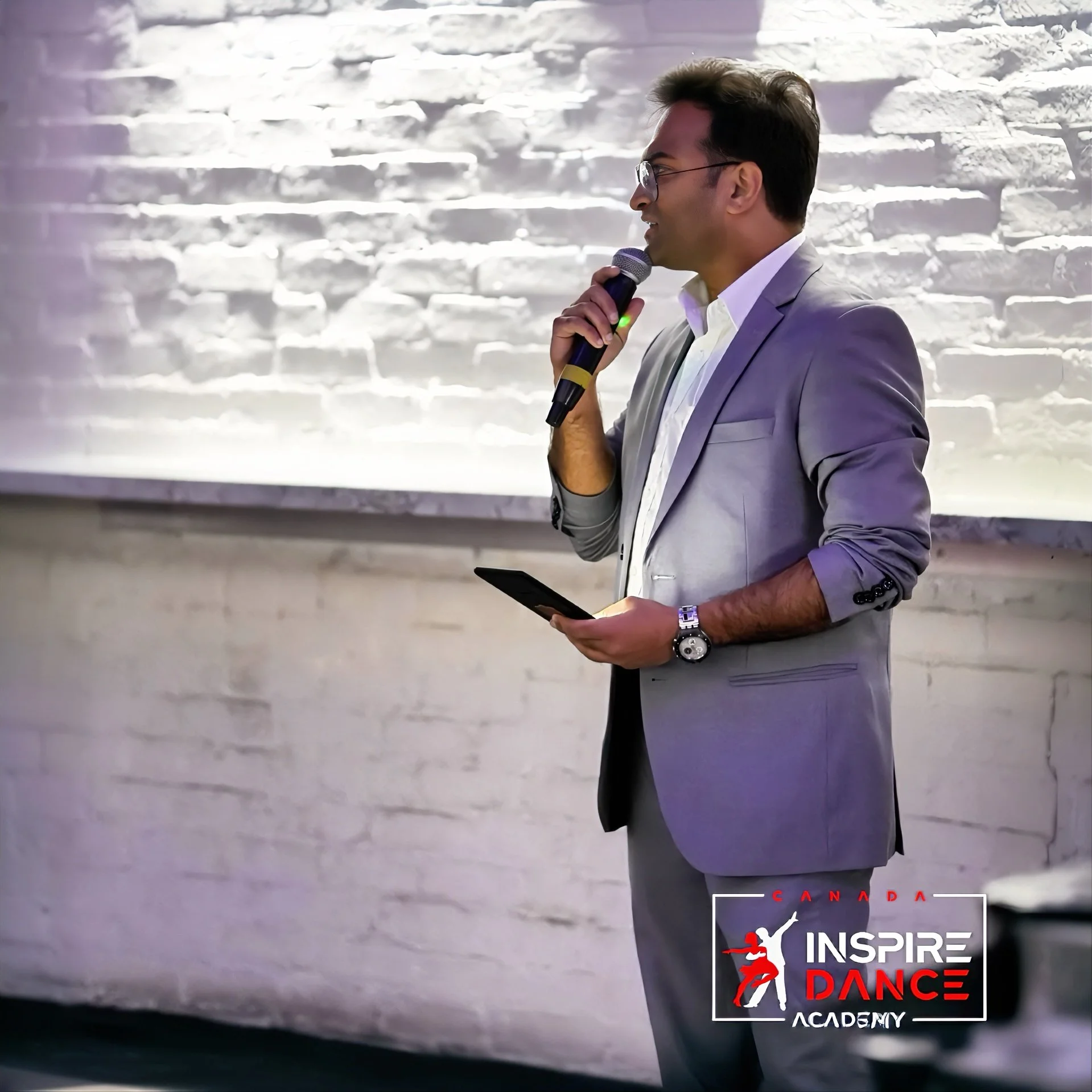A man in a gray suit and glasses speaking into a microphone at an Inspire Dance Academy event, standing in front of a white brick wall with a logo of the academy in the corner.