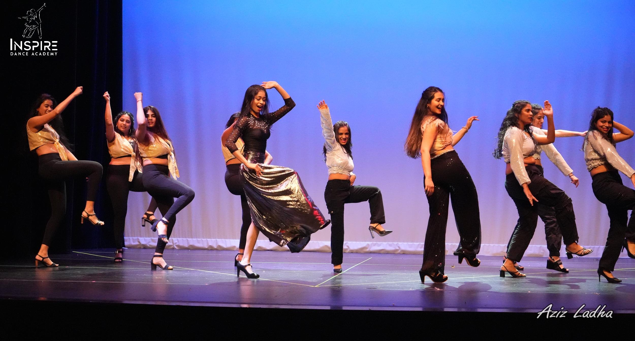 A group of young women performing a dance on stage, wearing black and gold outfits, with some raising their legs and smiling, under stage lights with a blue backdrop. The Inspire Dance Academy logo is in the top left corner, and Aziz Ladha is credited in the bottom right corner.