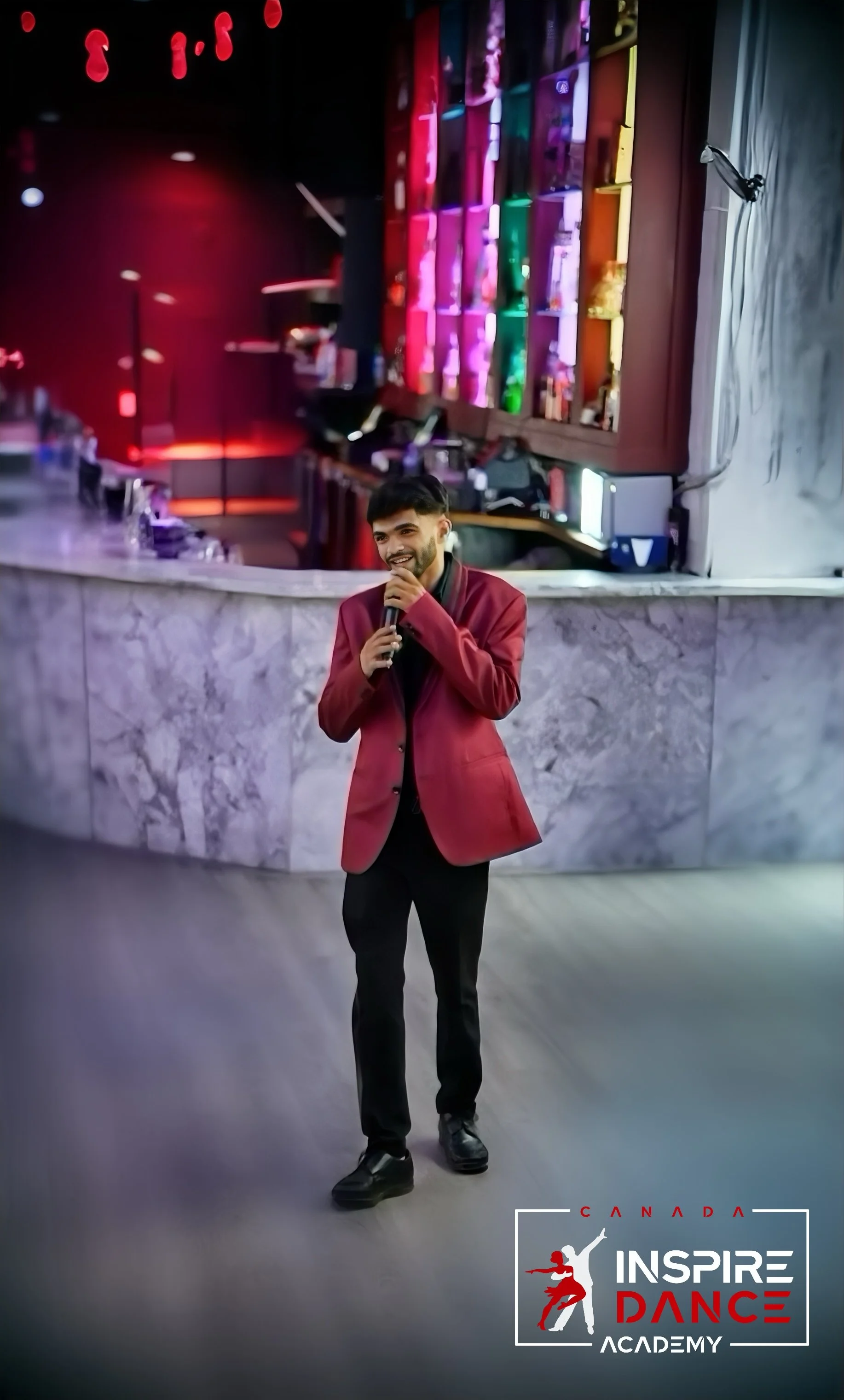 A man in a red blazer holding a microphone, smiling, standing in front of a colorful stage with 'Canada Inspire Dance Academy' logo in the bottom right corner.