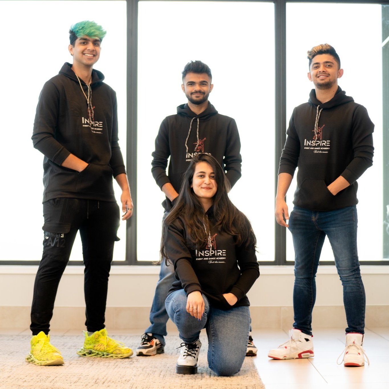 Group of four young adults wearing black hoodies with 'INSPIRE' logo, standing on a carpeted floor in front of large windows. One woman kneeling in front, smiling, three men standing behind her.