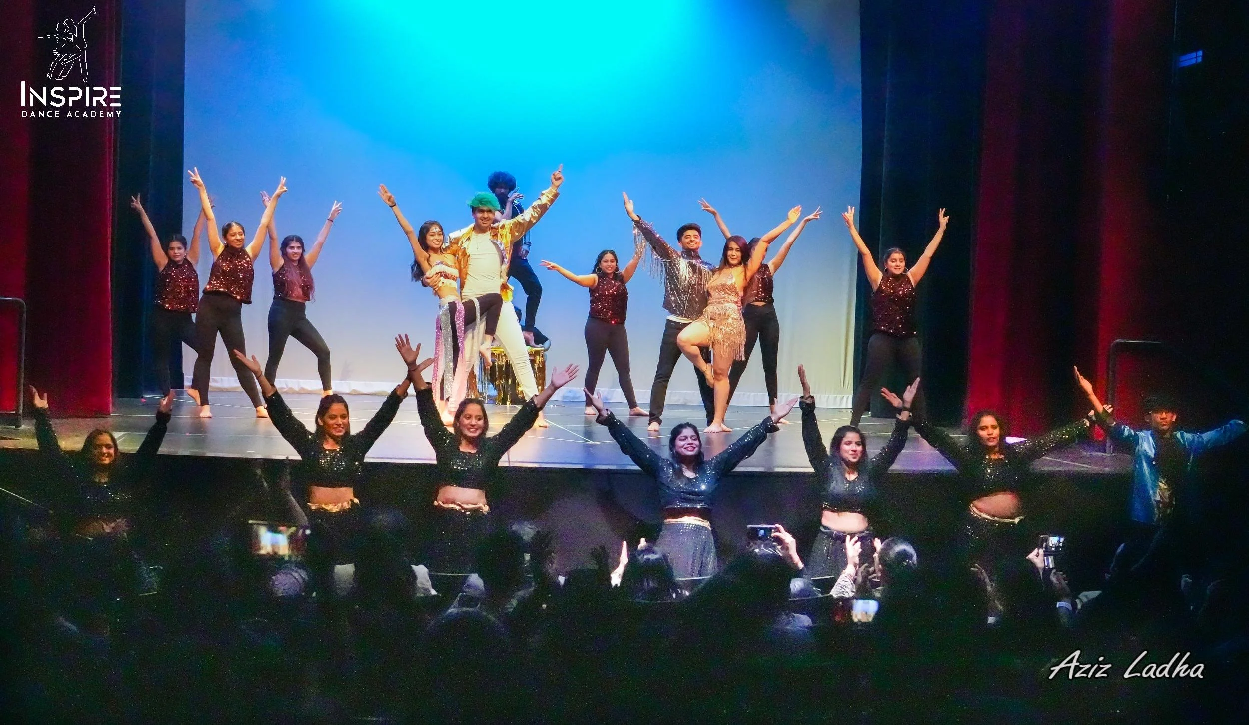 Stage with dancers performing, wearing colorful costumes, audience watching, and a backdrop with the Inspire Dance Academy logo.