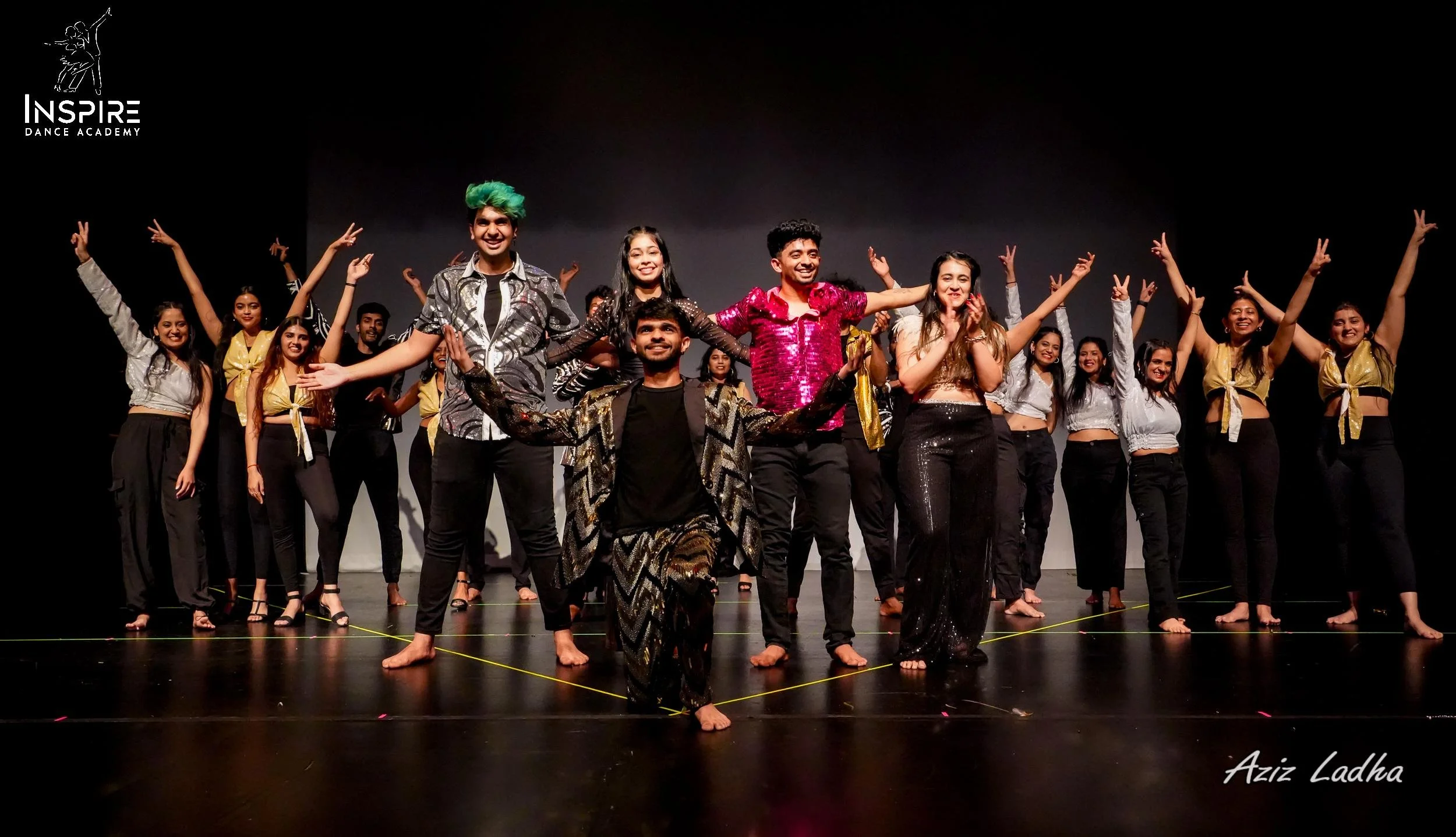 Group of dancers on stage performing, with some in colorful costumes and others in casual attire, smiling and raising their arms. Black background, stage lights, Inspire Dance Academy logo in top left corner, and Aziz Ladha's signature in bottom right corner.