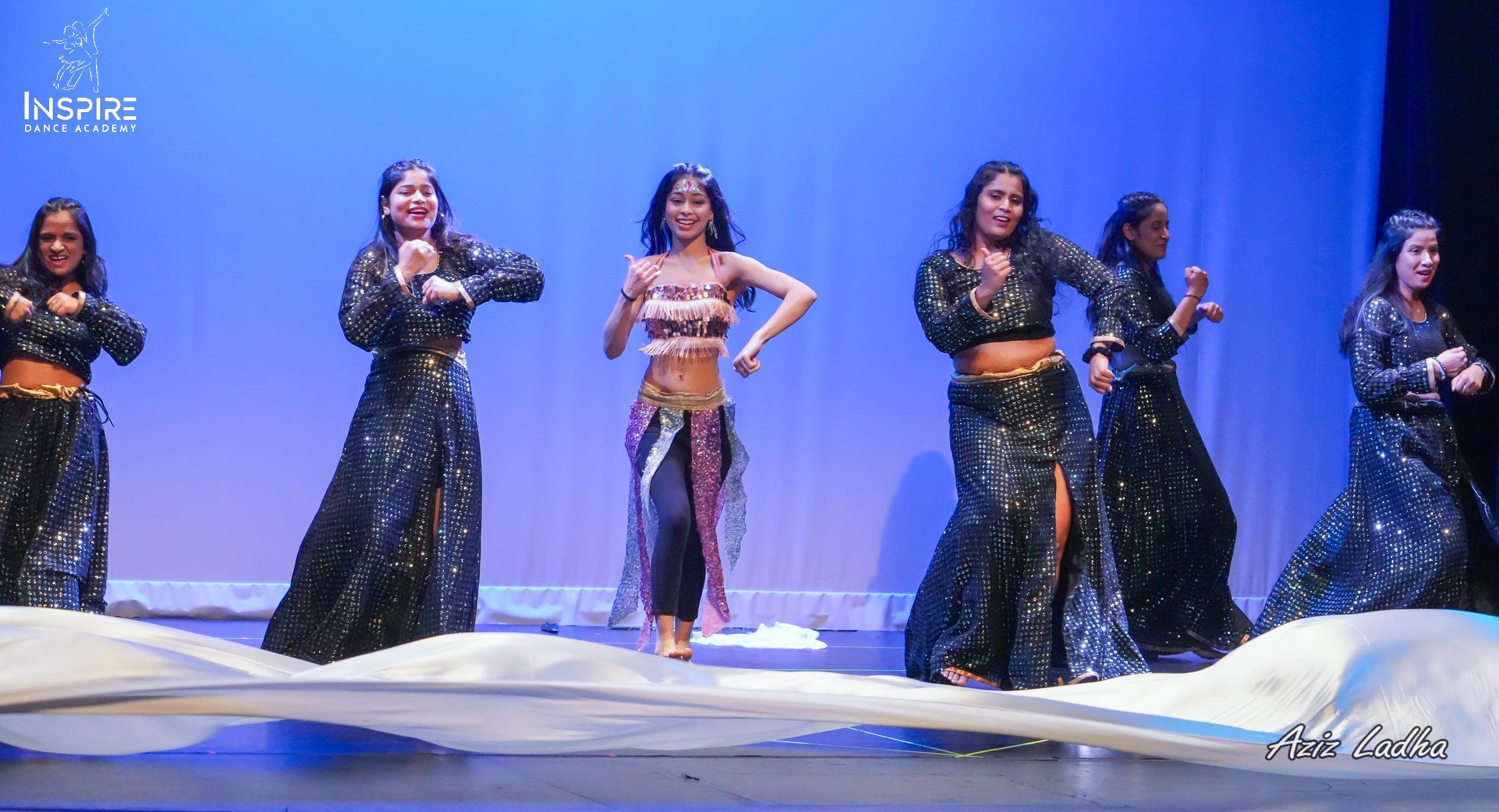 Group of six young women performing a dance on stage with a blue backdrop. They are dressed in black, shiny, sequined outfits, and one woman in the center wears a colorful costume with fringe and sequins.