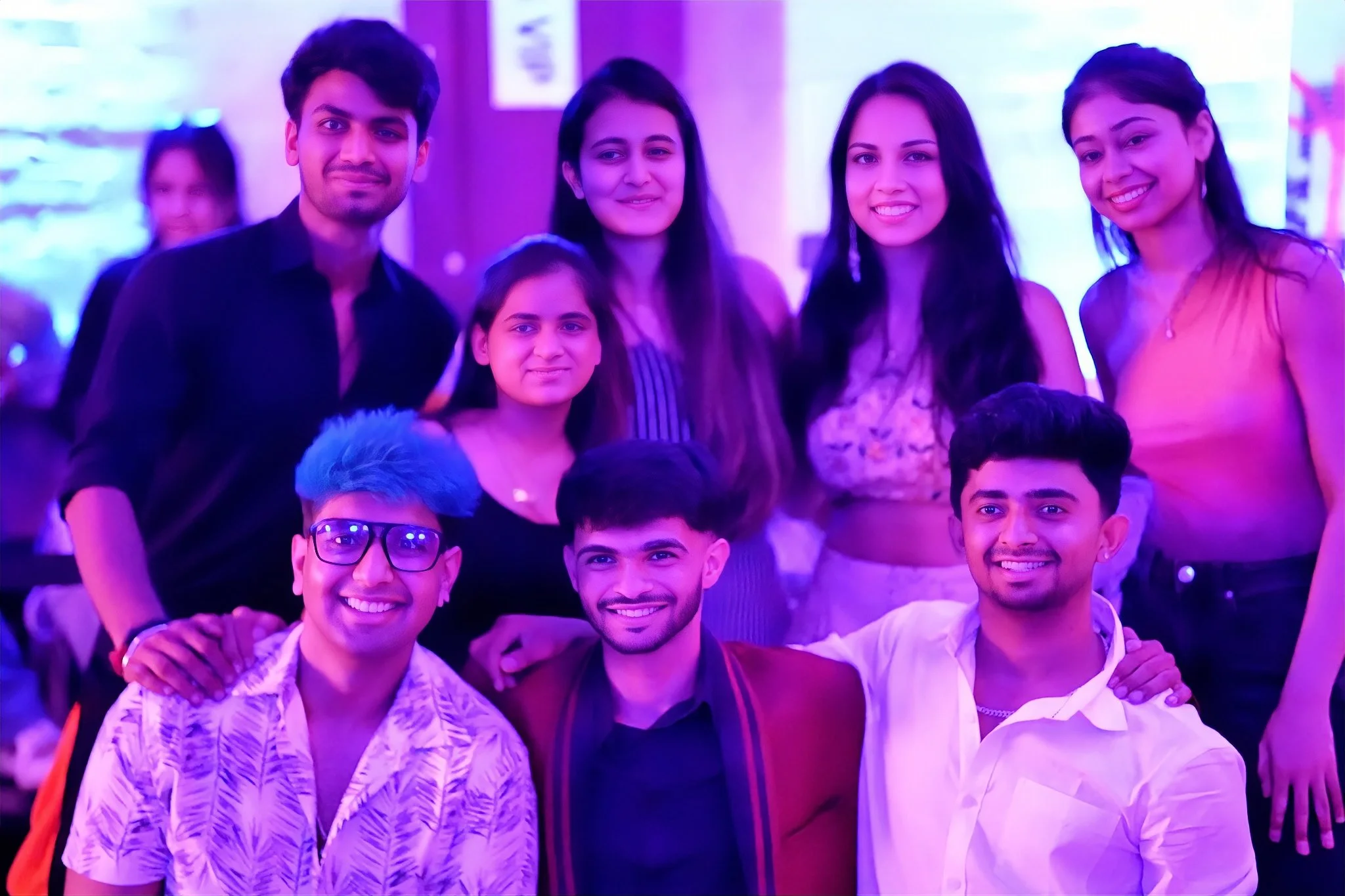 A group of eight young adults, five women and three men, smiling and posing together at a party or event with colorful lighting.