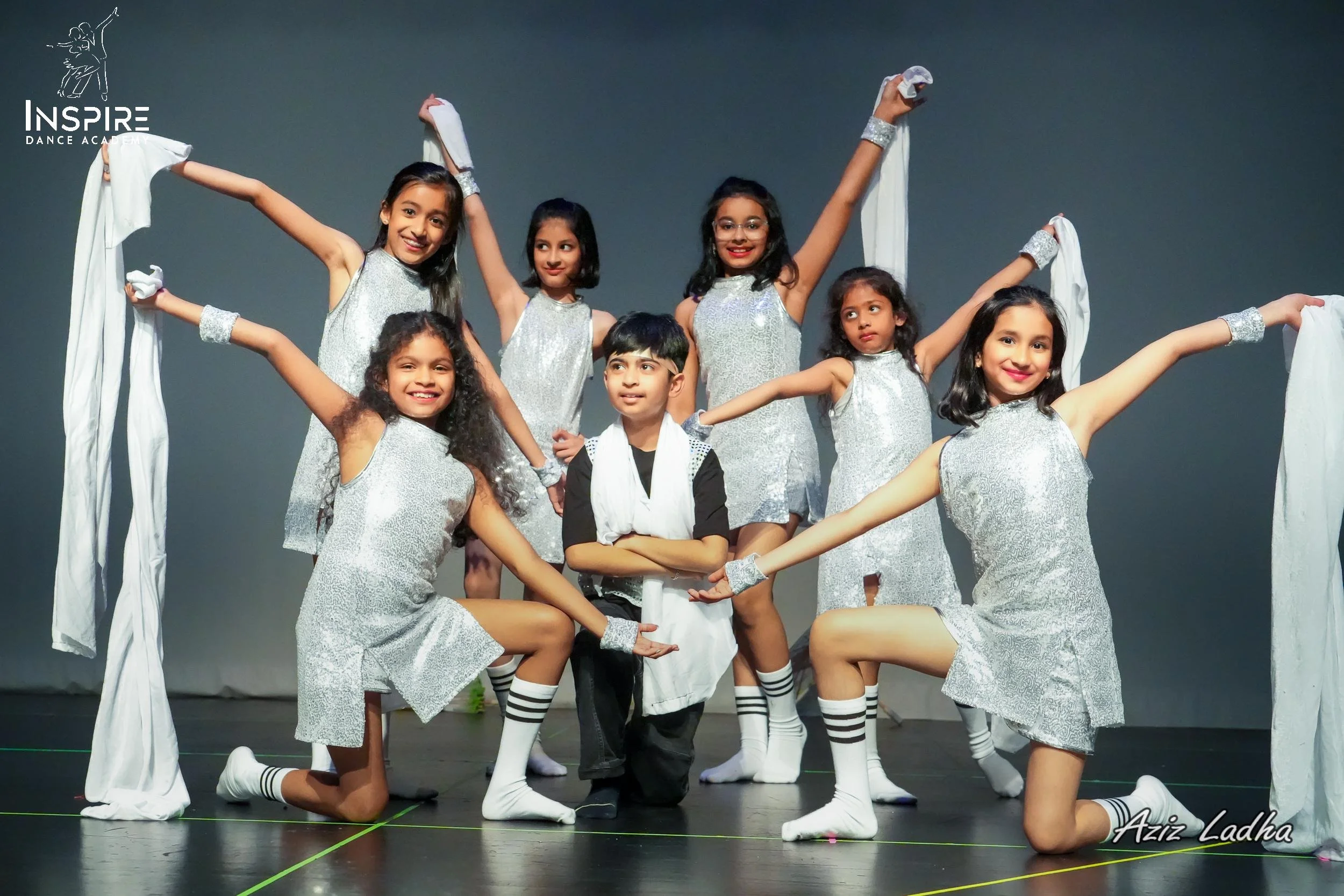 Group of young children dressed in silver costumes posing on stage during a dance performance, with some kneeling and one child standing in the center. The background is plain gray.