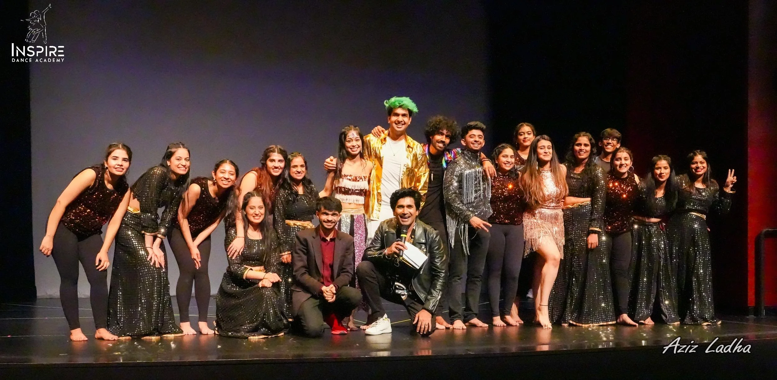 Group of dance performers on stage, some in black sparkly costumes, others in colorful costumes, posing and smiling after a performance, with a dark stage background, and the Inspire Dance Academy logo in the top left corner.