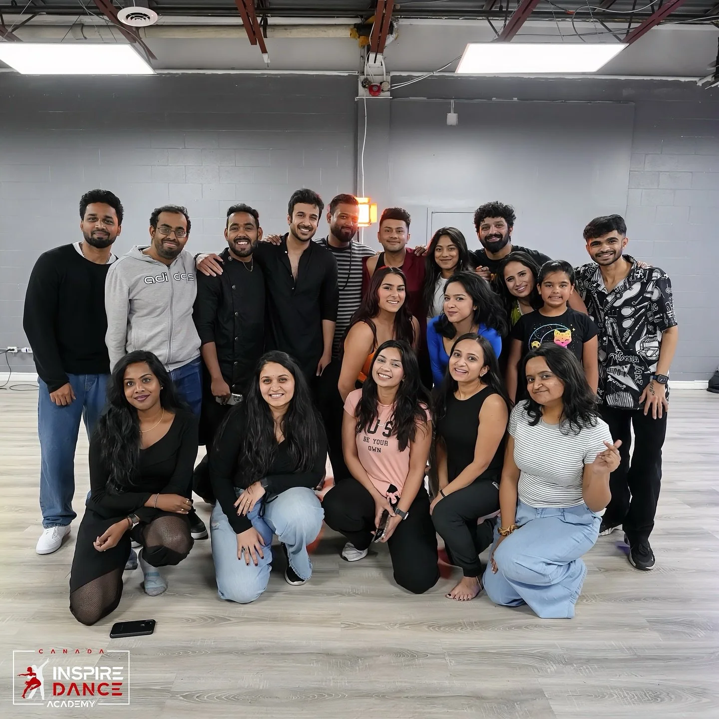 Group of 16 people posing together in a dance studio with gray walls and a wooden floor, including a mix of men and women, some smiling and some serious, some kneeling and some standing, with