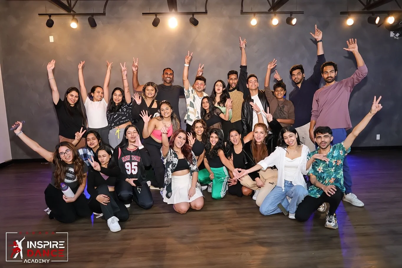 Group of young people smiling and posing for a photo in a dance studio with a dark gray wall and wooden floor. Some are standing, others kneeling, with many raising their hands. The Inspire Dance Academy logo is in the bottom left corner.