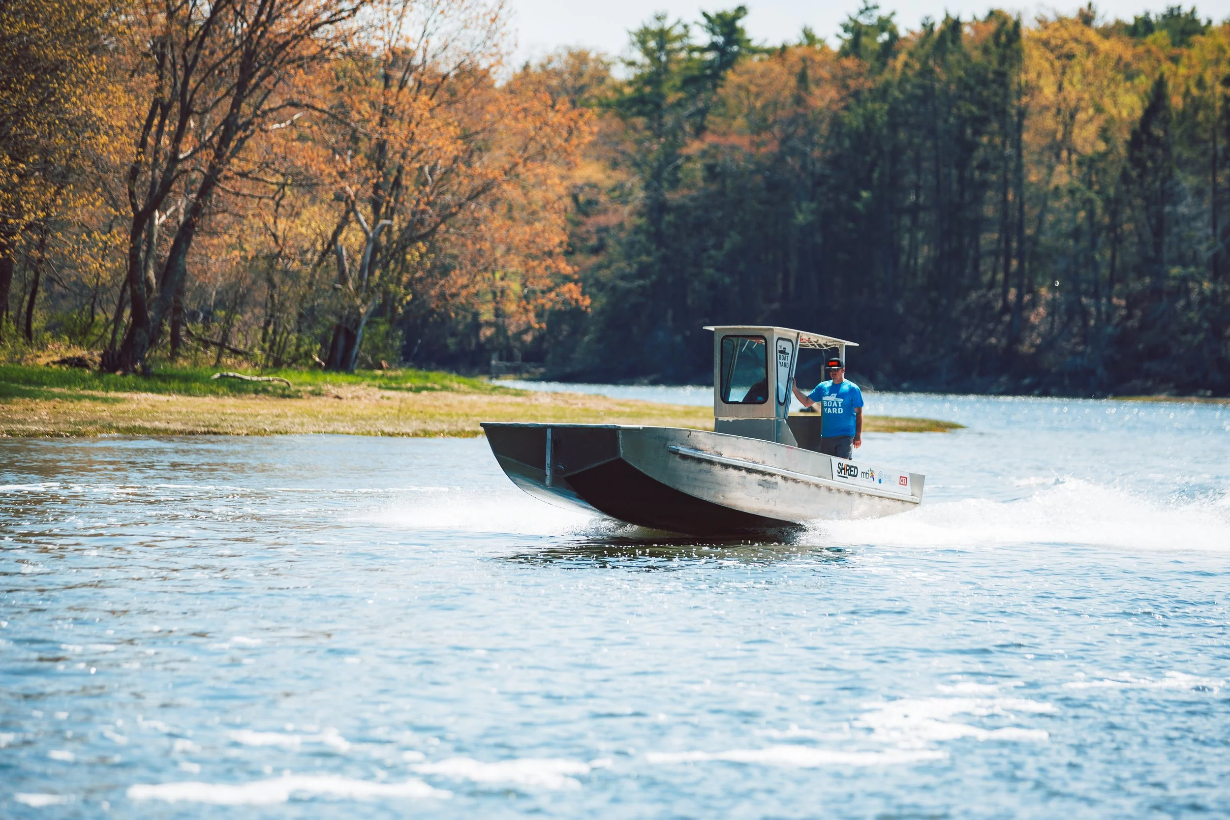 A man driving a small boat on a river with autumn trees in the background.
