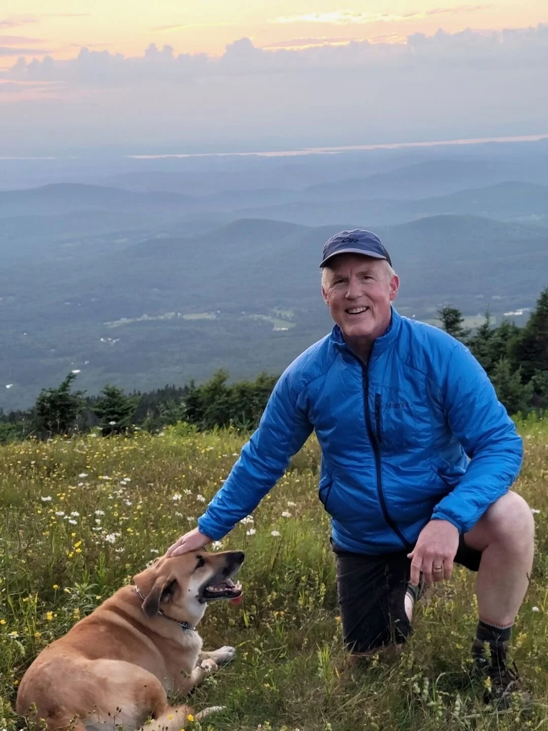 Brian Degen in a blue jacket in a field with flowers and mountain views with his dog Lilah.