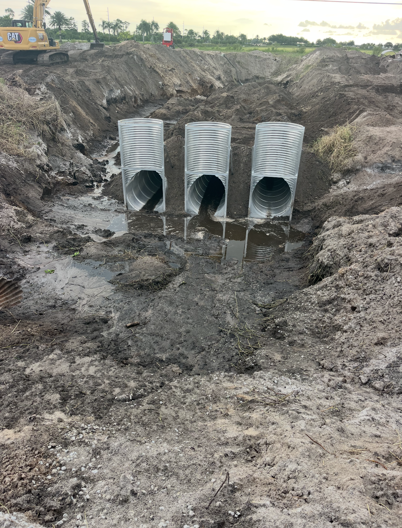 Three metallic underground pipes installed in a trench, with muddy water surrounding them, at a construction site with excavators and soil mounds in the background.
