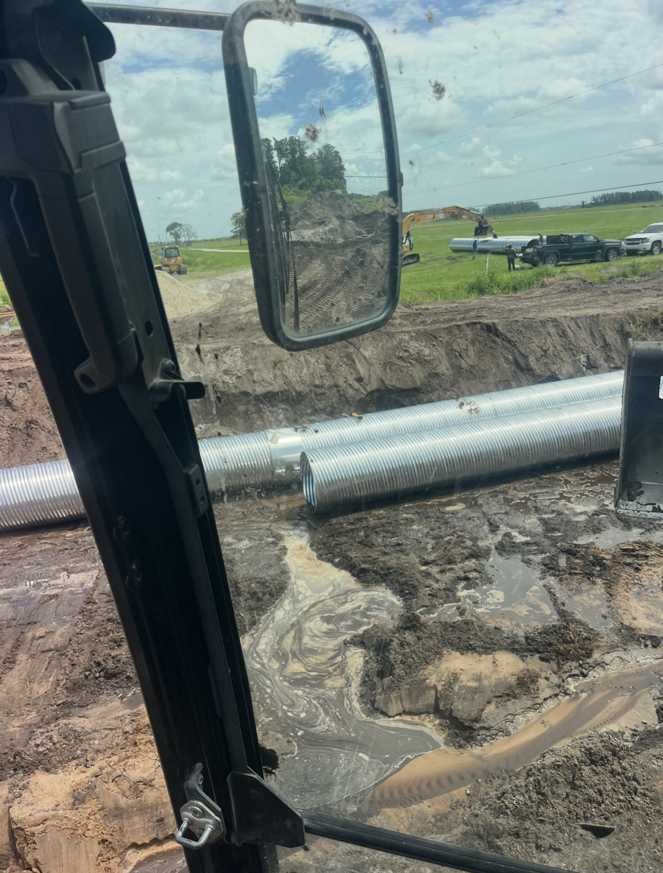 View from inside a construction vehicle showing large metallic pipes on muddy ground and people working on construction site in the background under partly cloudy sky.