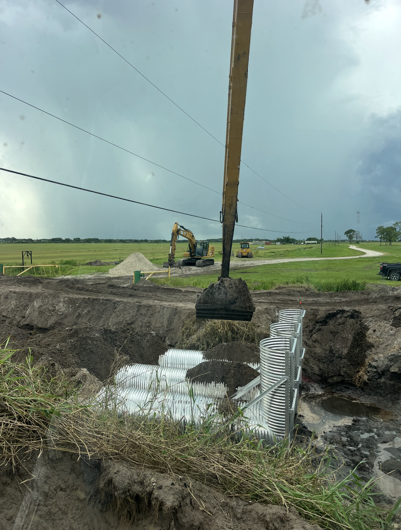 Construction site with large yellow machinery, a fallen utility pole with wires, underground pipe installation, and a dirt road in a rural area under cloudy sky.
