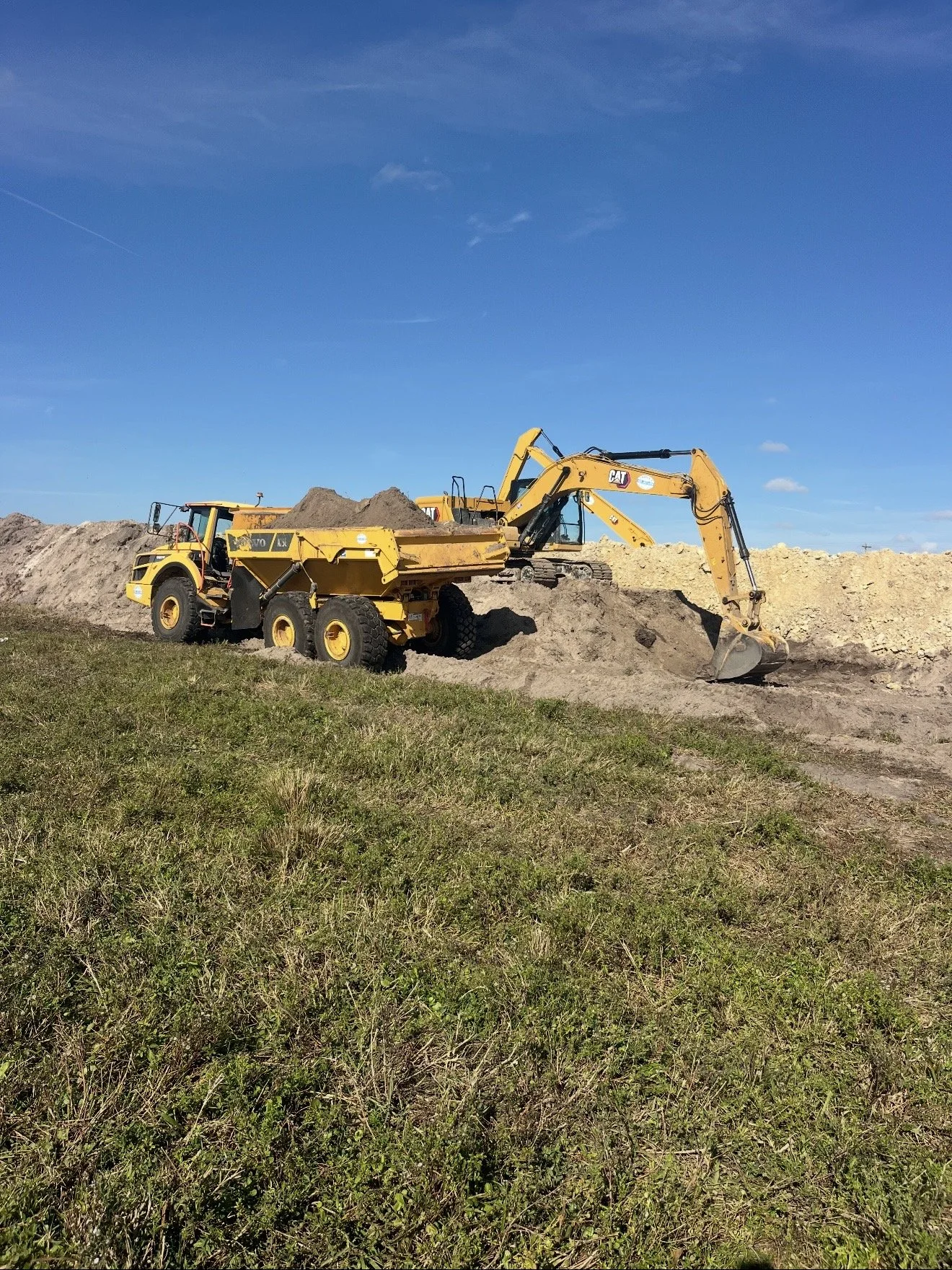 A yellow construction vehicle, a dump truck, is parked on grass with an excavator digging up dirt nearby, under a blue sky with a few clouds.