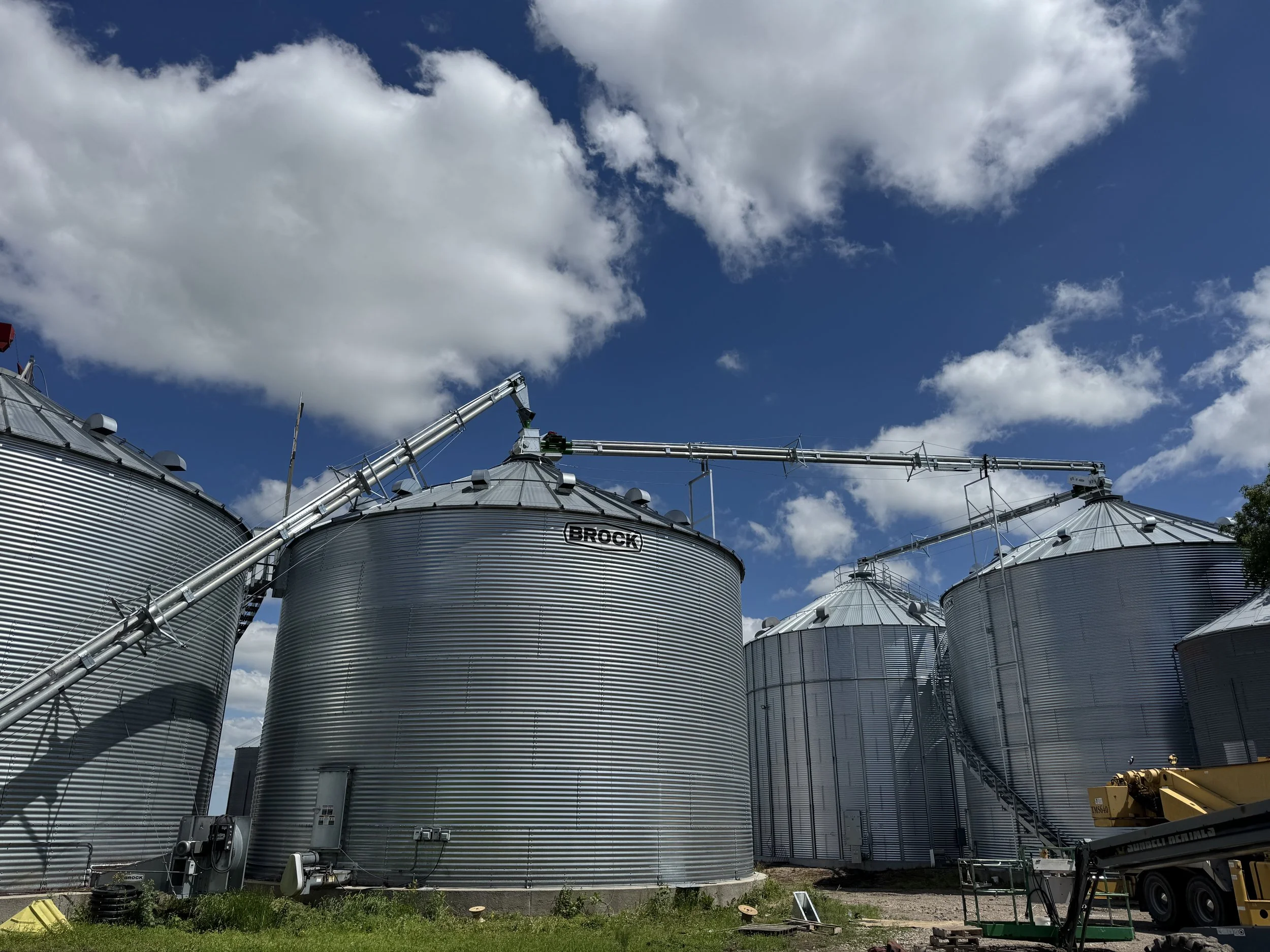 Several large metal grain silos with a clear blue sky and scattered clouds above, connected by metal pipes.