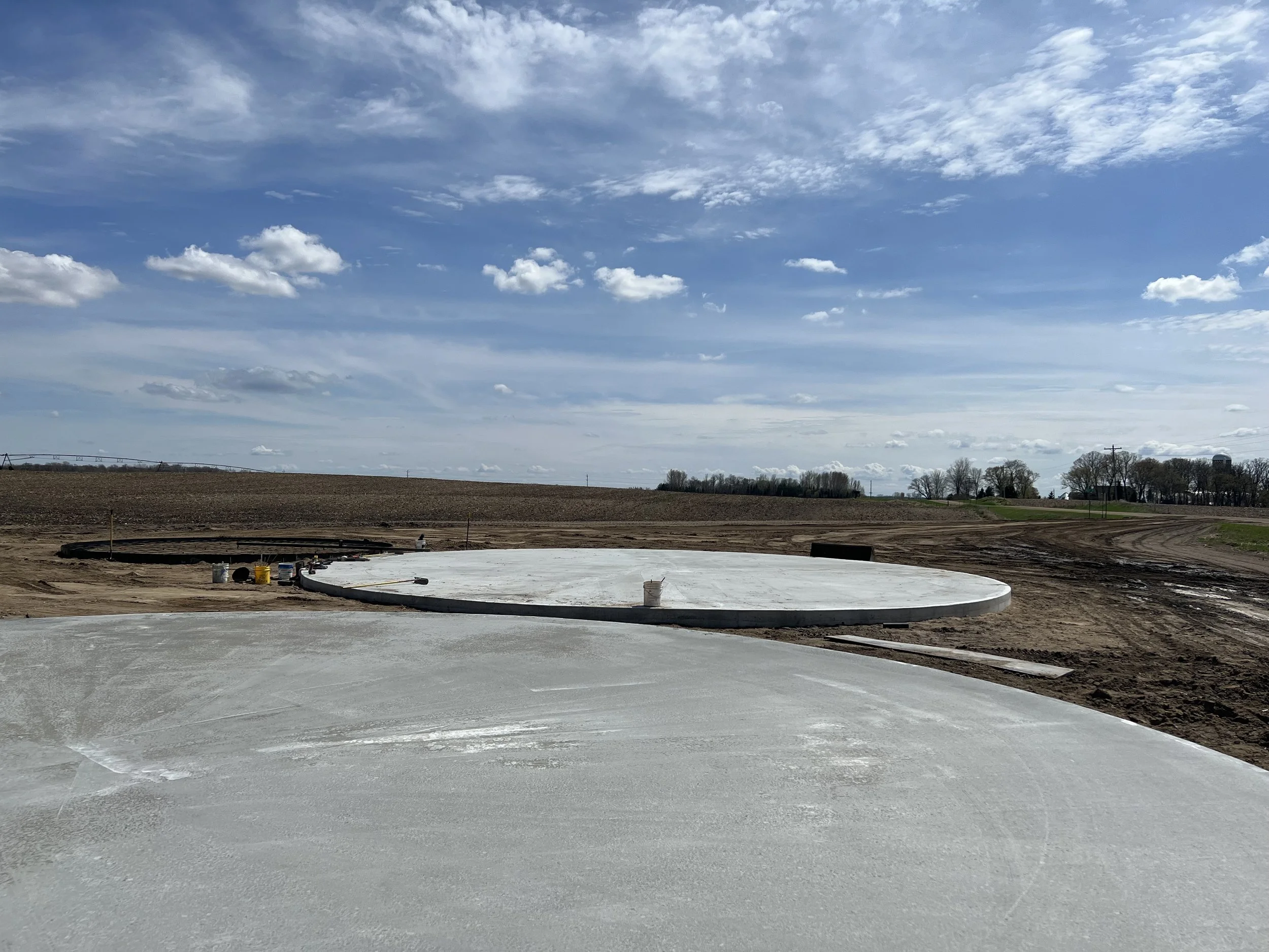 Construction site with circular concrete foundation on a farm under a partly cloudy sky.