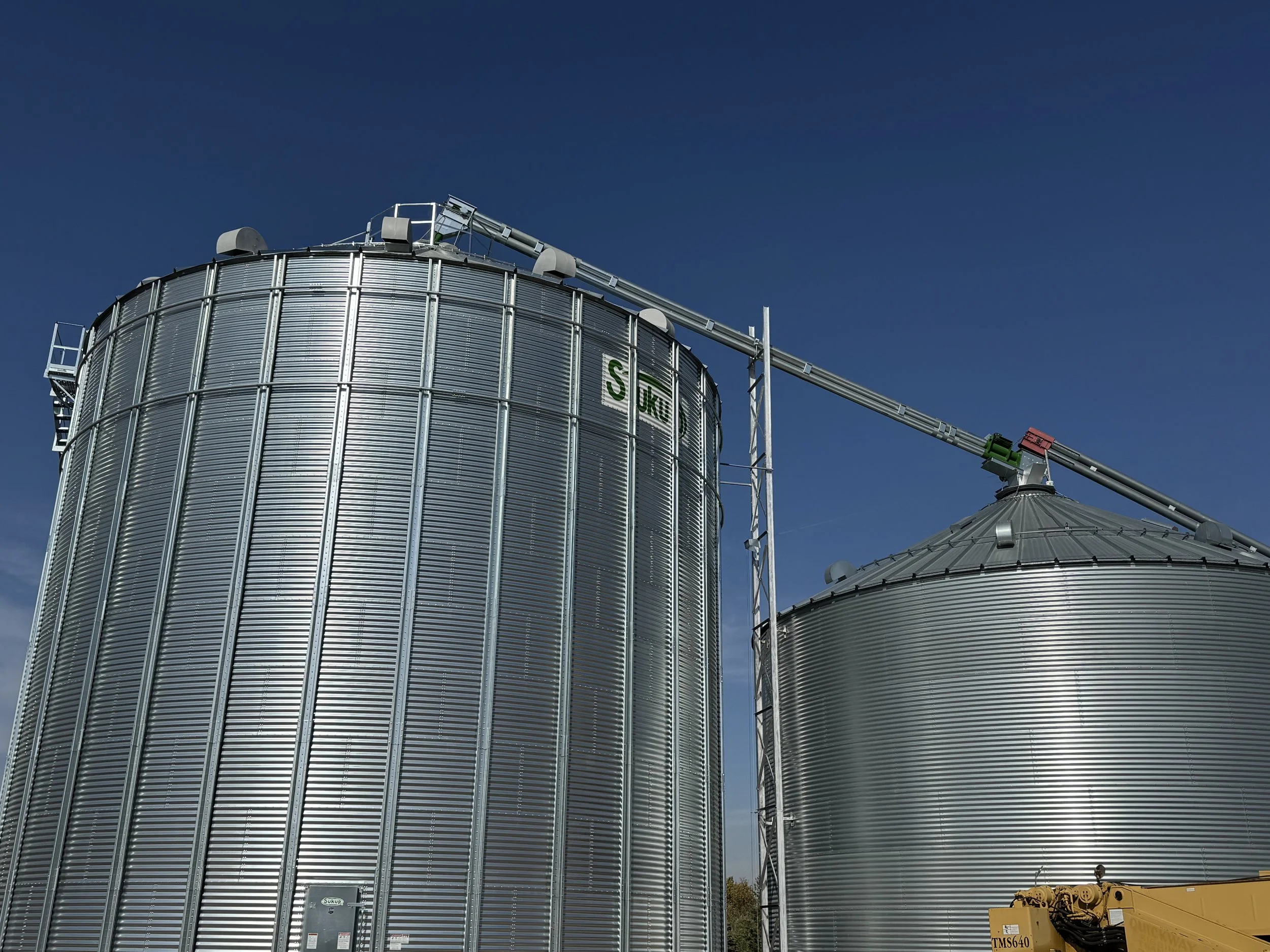 Two large cylindrical metal grain silos with a conveyor system connecting them, set against a clear blue sky.