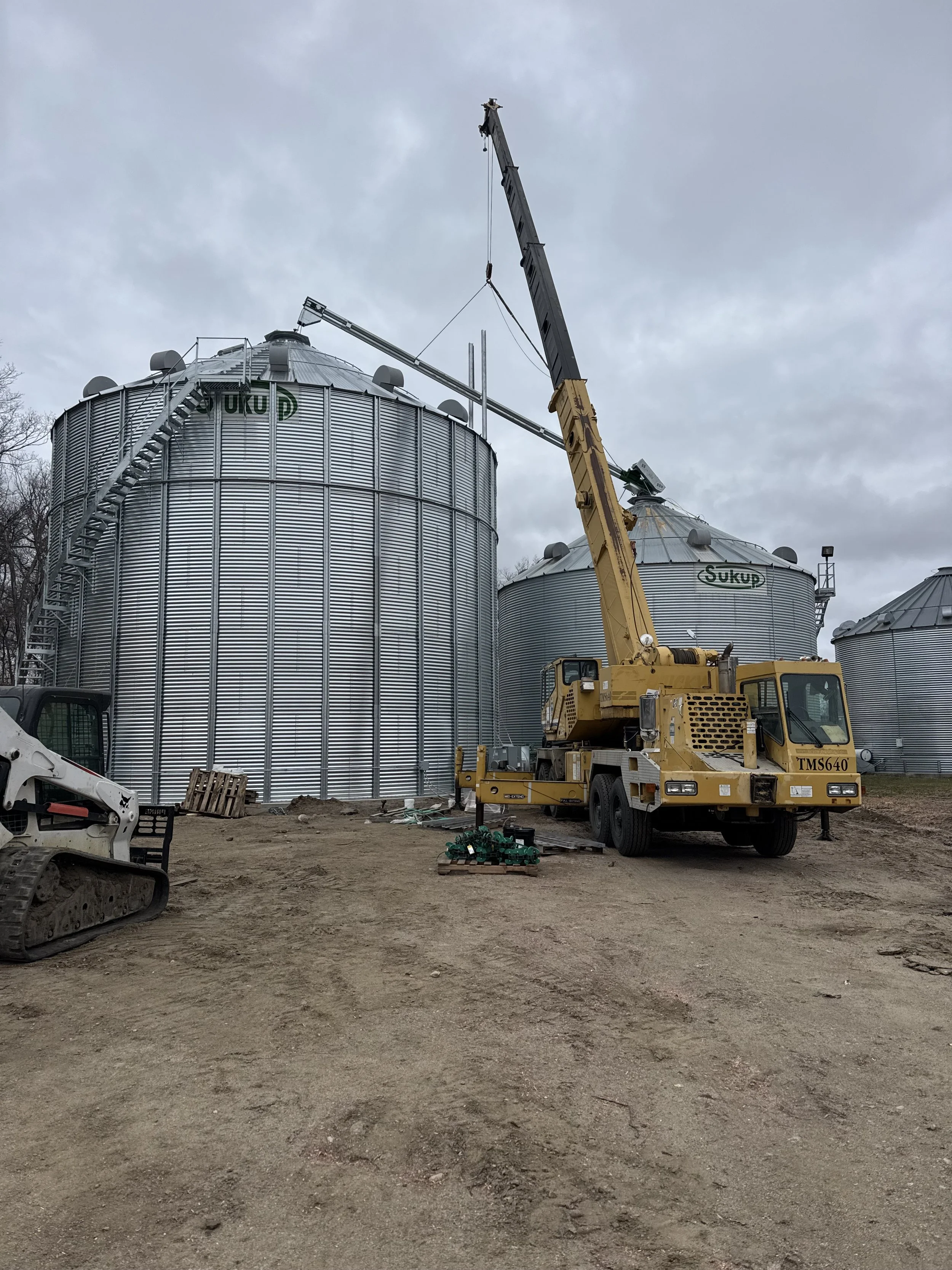A large yellow crane is lifting a metal silo in an industrial area with more silos in the background and a cloudy sky overhead.