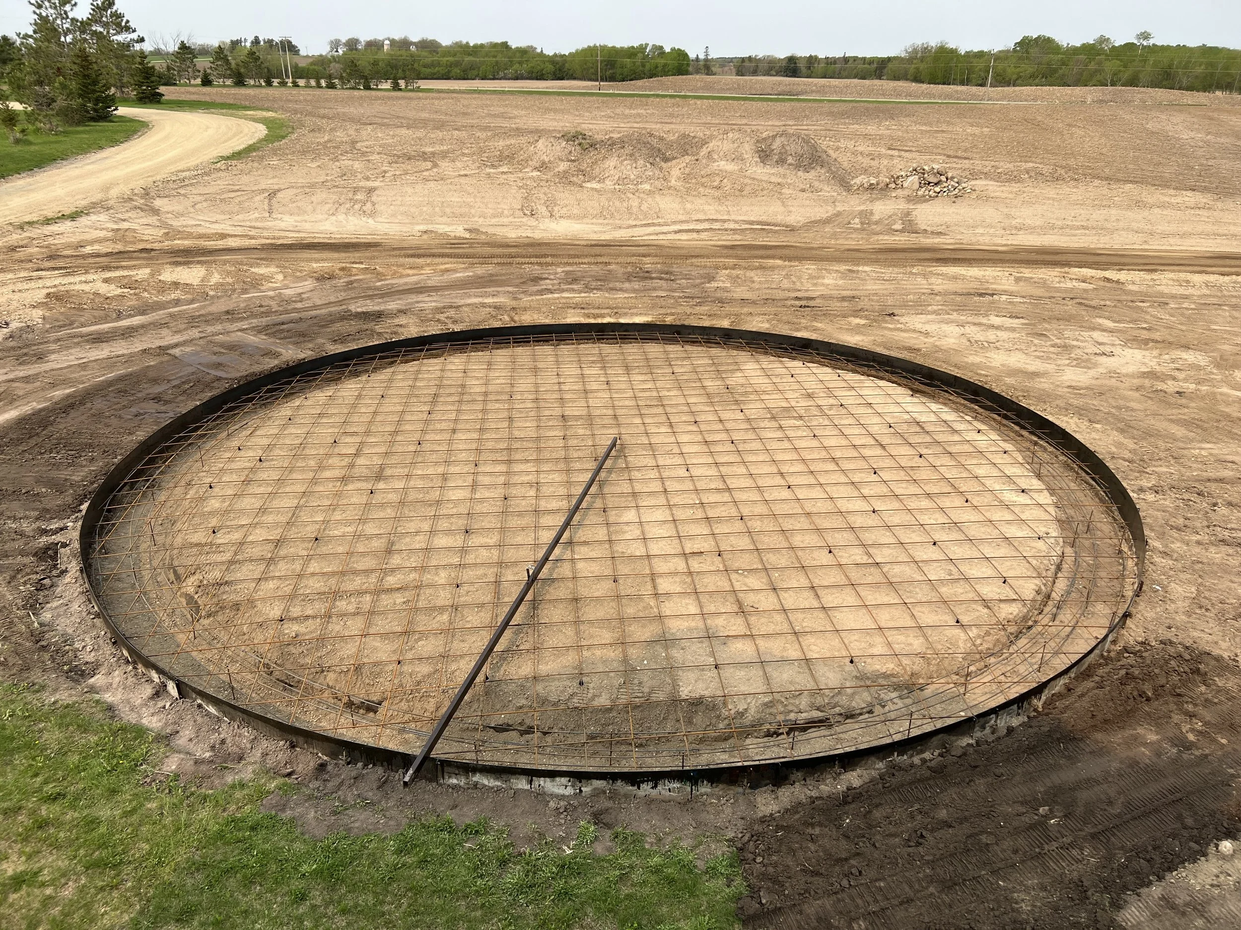 Construction site with a large circular hole reinforced with metal rebar grid and frame, surrounded by dirt and grass, with a dirt road in the background.