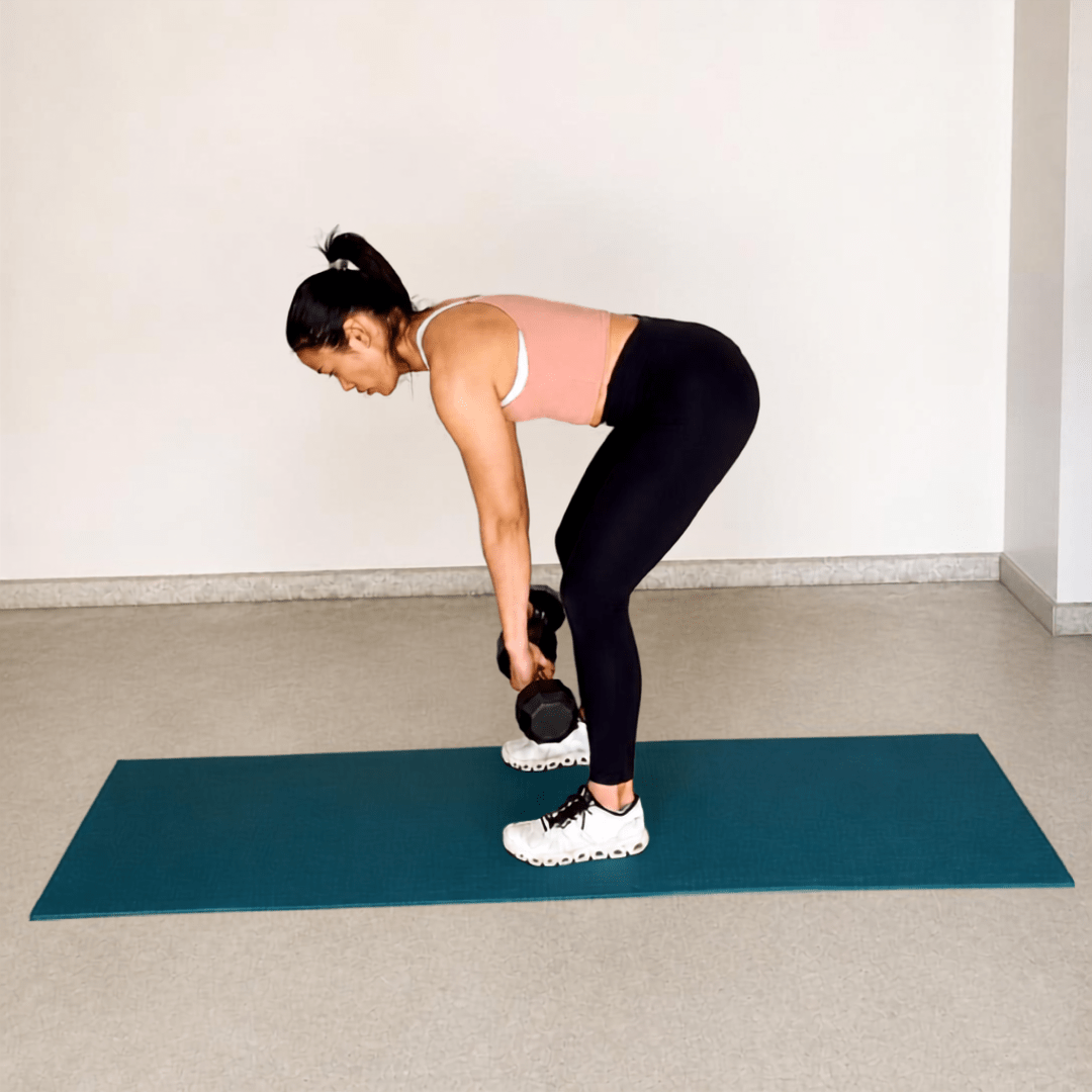 Woman doing a dumbbell deadlift on a mat, in a pink top and black leggings, demonstrating hamstring strength.