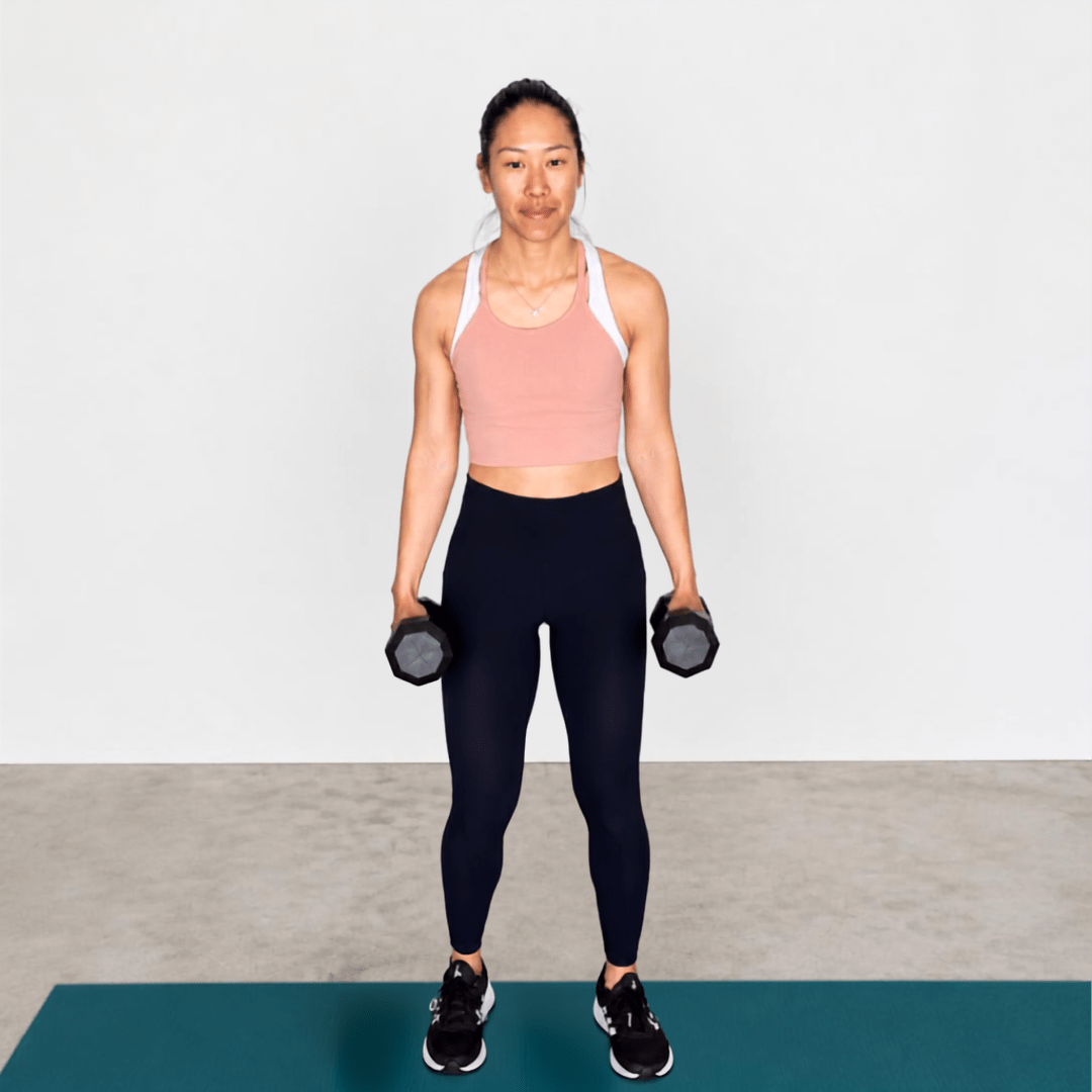 Woman doing a Farmer’s Carry on a teal mat, in a pink top & black leggings, holding dumbbells to show grip and core strength