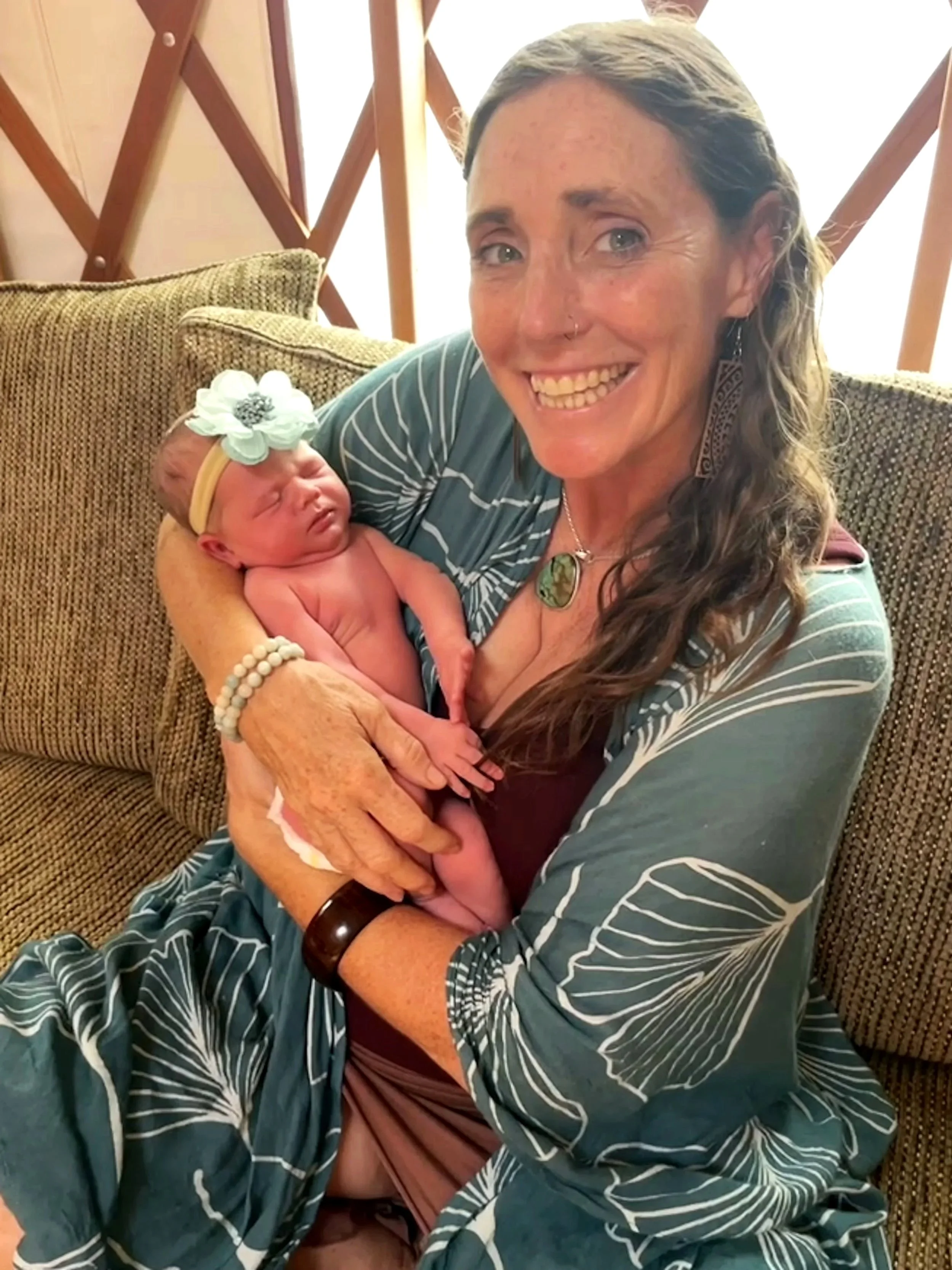 Hope Medford,  birth keeper, midwife, doula, and founder of the sacred birth path, smiling while holding a newborn baby who was born healthy, at home, a few days earlier. The baby is wearing a sweet flower headband.  SacredBirthPath.com