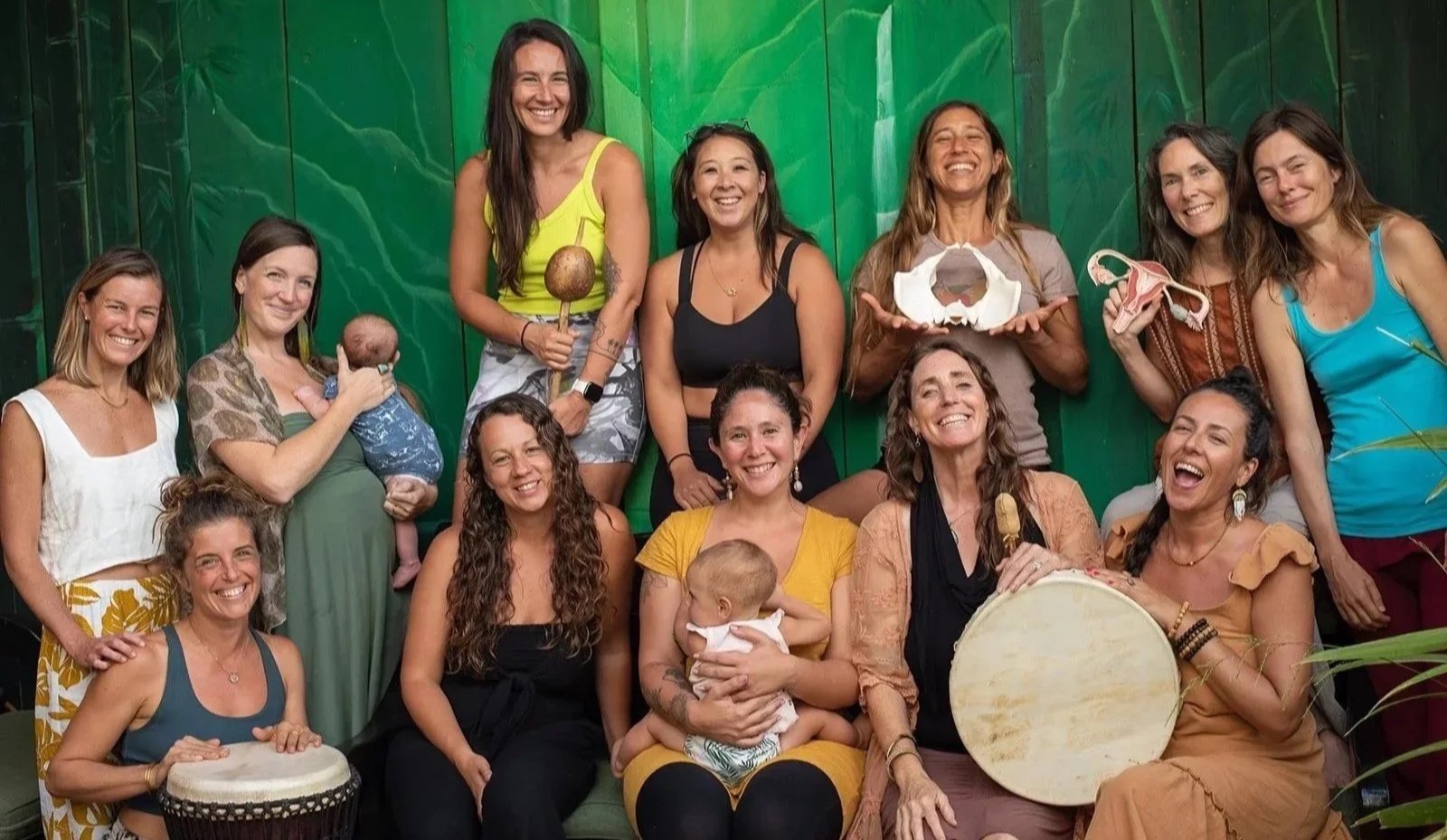 Sacred Birth Class in Maui of doulas, birth keepers, students, mamas, babies and their teacher, Hope Medford, smiling and holding a drum, shaker, a pelvis, and a uterus model, and enjoying the sisterhood of sacred birth work.  SacredBirthPath.com