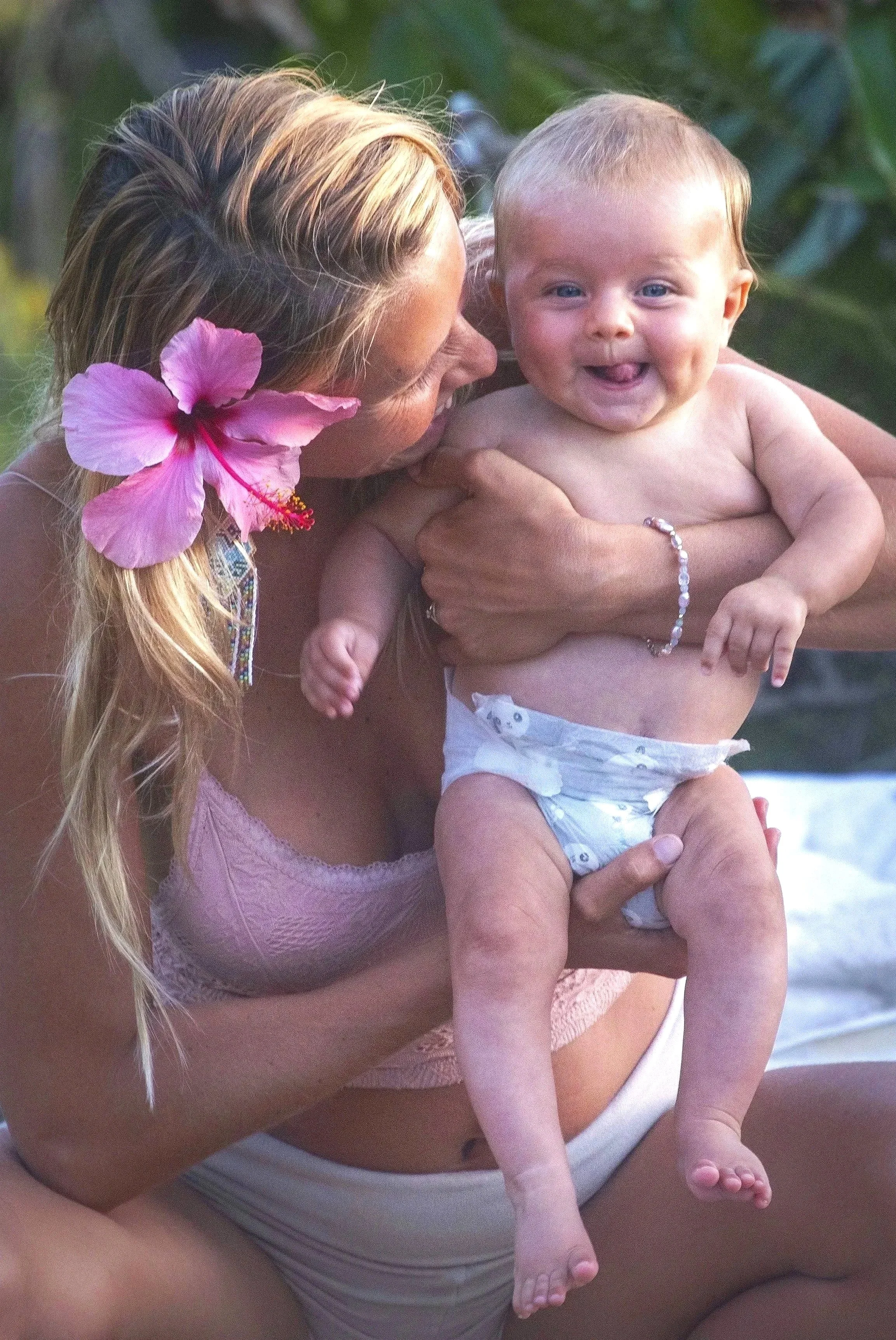 A beautiful woman with a pink hibiscus flower in her hair is holding her sweet, happy baby outdoors during a holistic doula training in Hawaii.  SacredBirthPath.com